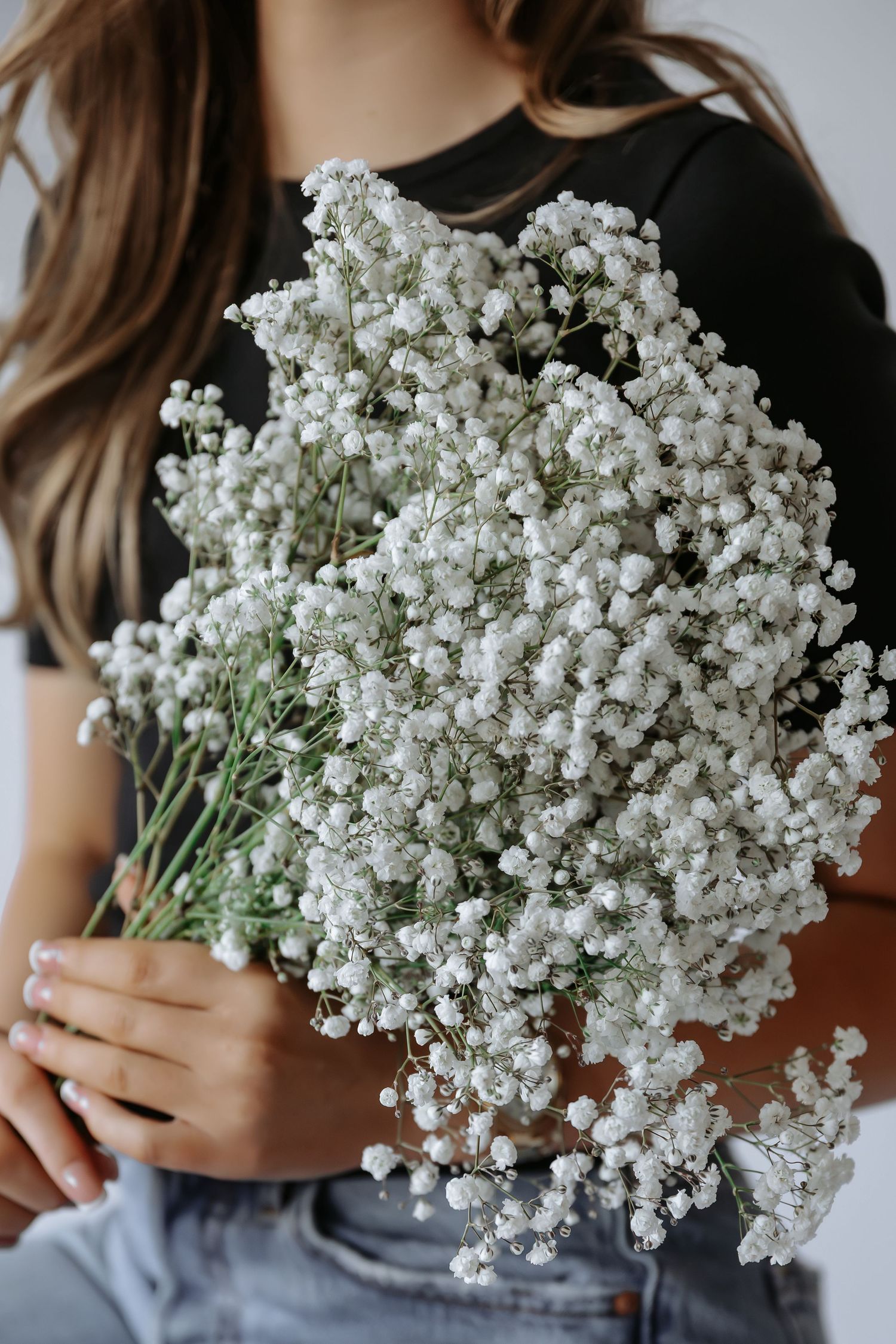 Woman in black top and jeans holding large bouquet of white baby's breath flowers against white background.