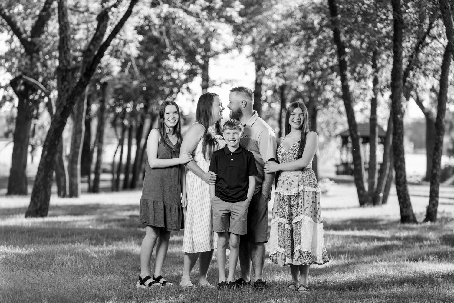 Black and white family portrait of five people posing together in a tree-lined park on a sunny day.