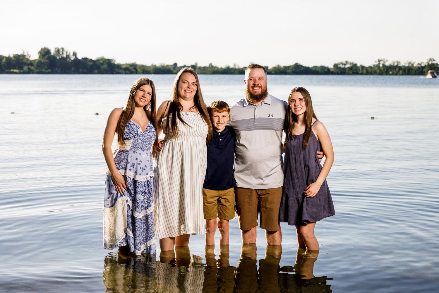 Smiling family of five standing in shallow lake water during a warm summer evening.