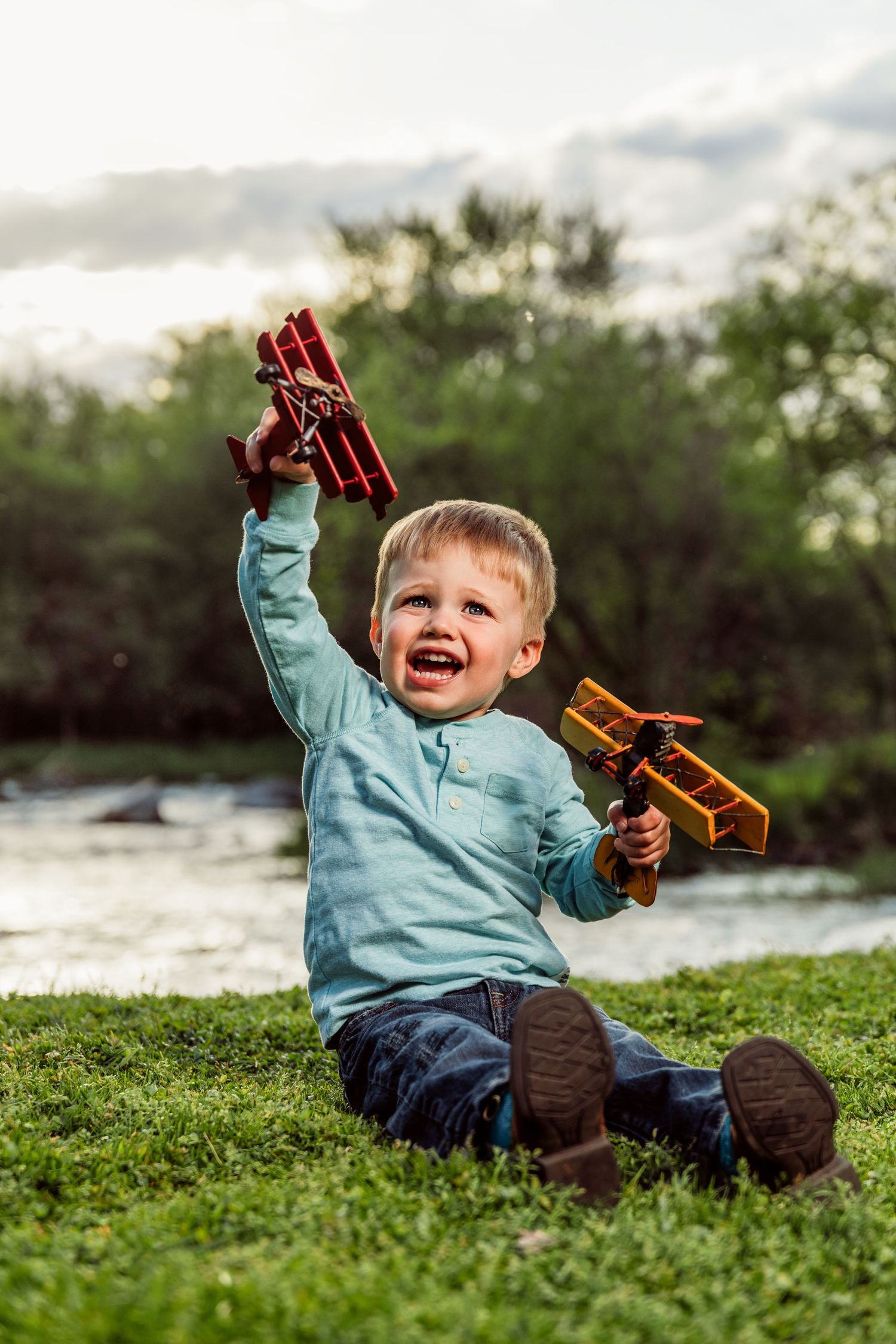A joyful toddler sits on grass holding two wooden toy airplanes up in the air outdoors near a river.