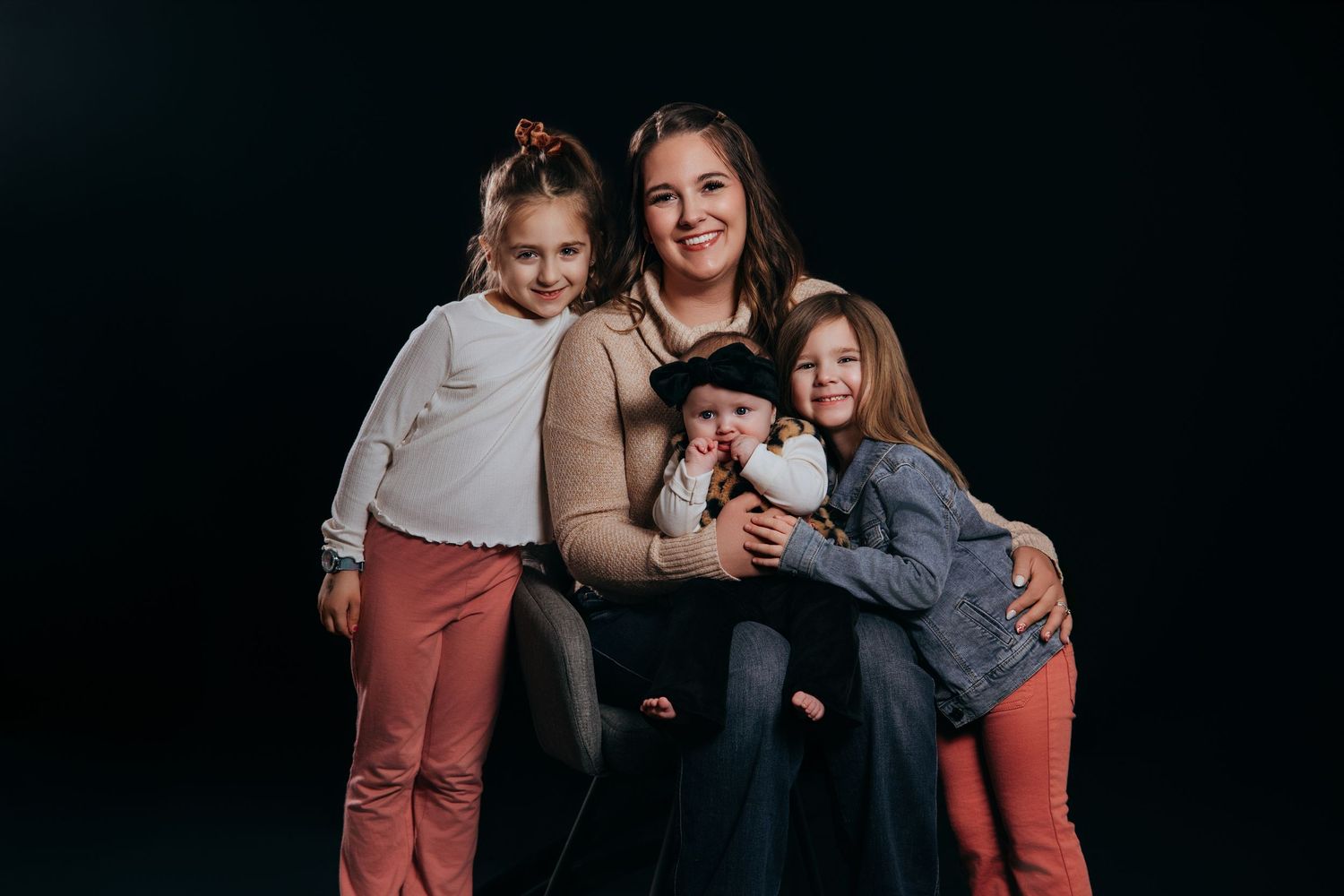 A mother sits with her three young daughters, all smiling, posed together against a dark studio background.