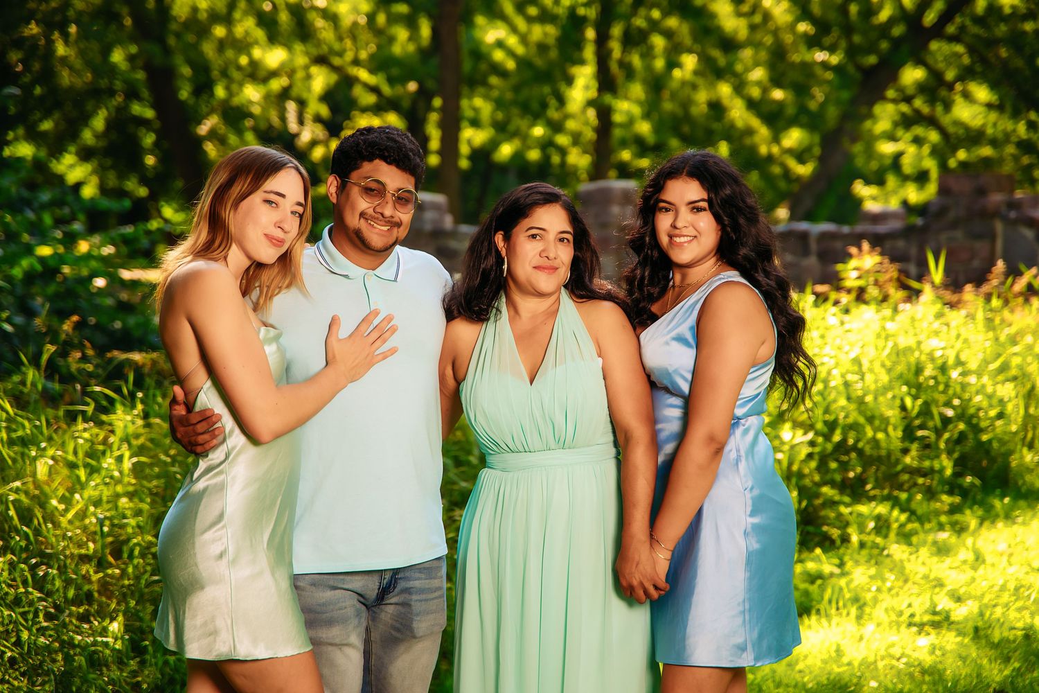 Four young adults posing together outdoors in a lush green park, wearing pastel outfits, smiling for a family portrait.