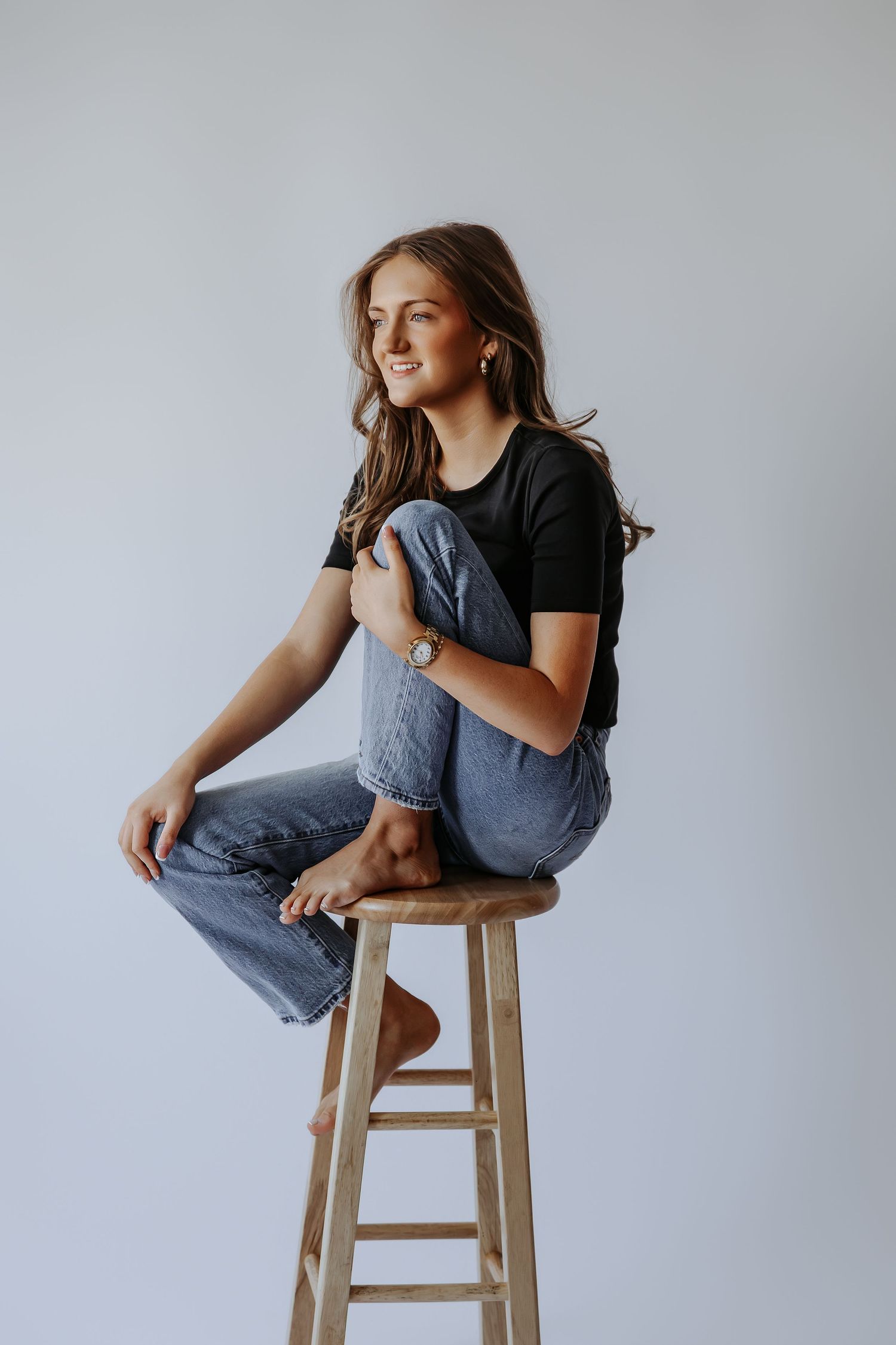 Smiling young woman in black t-shirt and jeans sitting barefoot on a wooden stool against a white background.