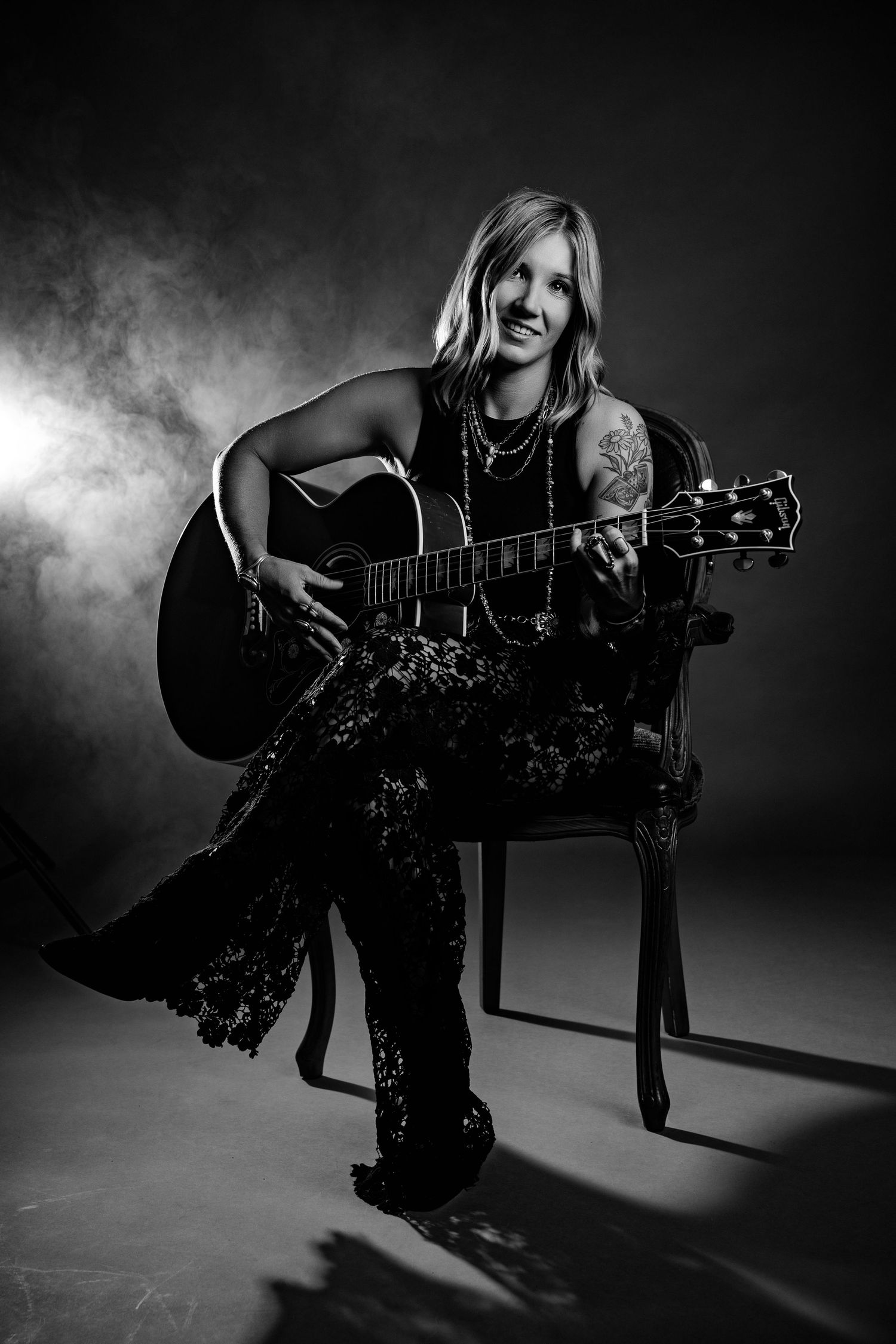 Black and white studio portrait of woman playing acoustic guitar seated in ornate chair.