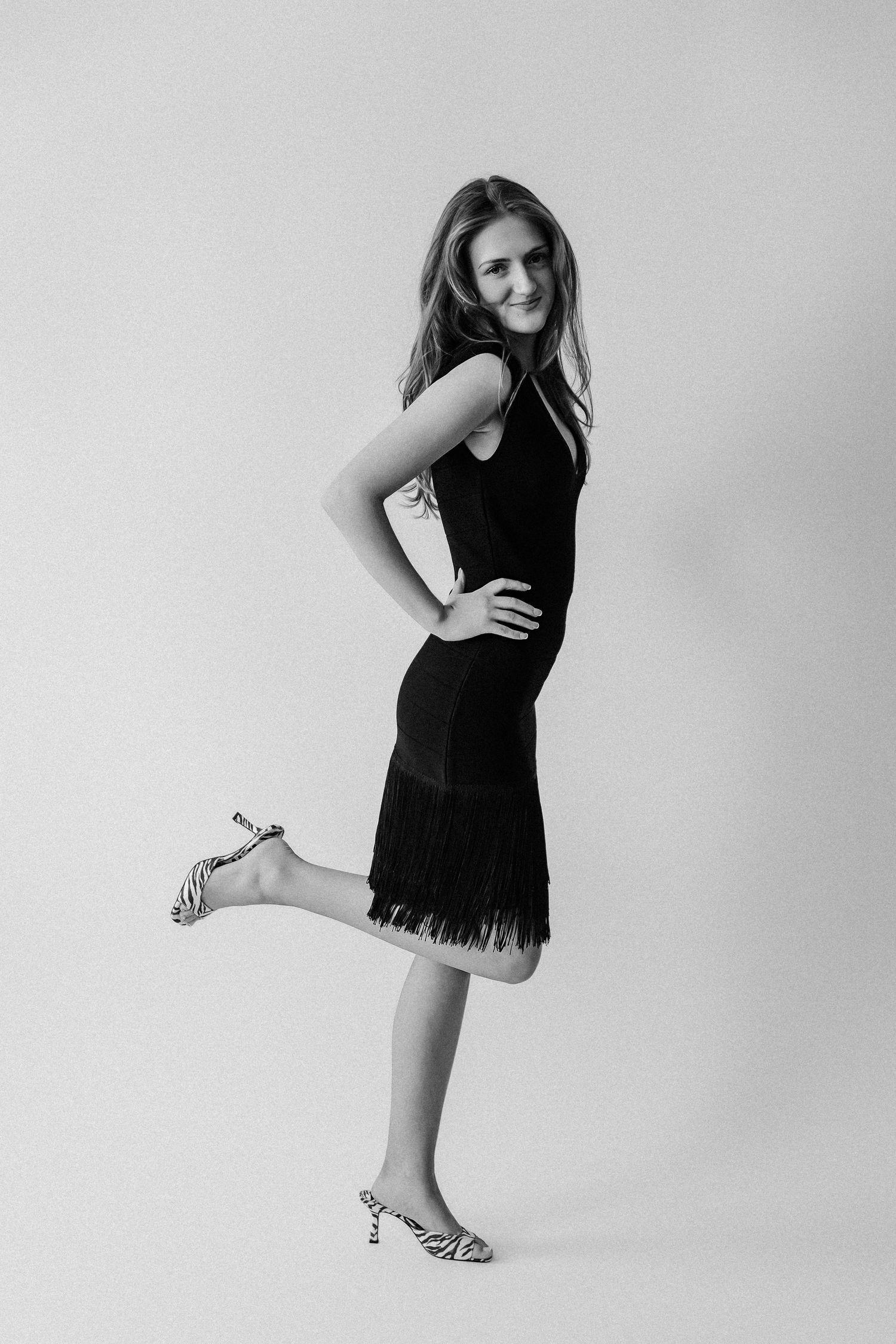 Young woman in black fringed dress and zebra heels posing playfully on one leg in black and white studio.