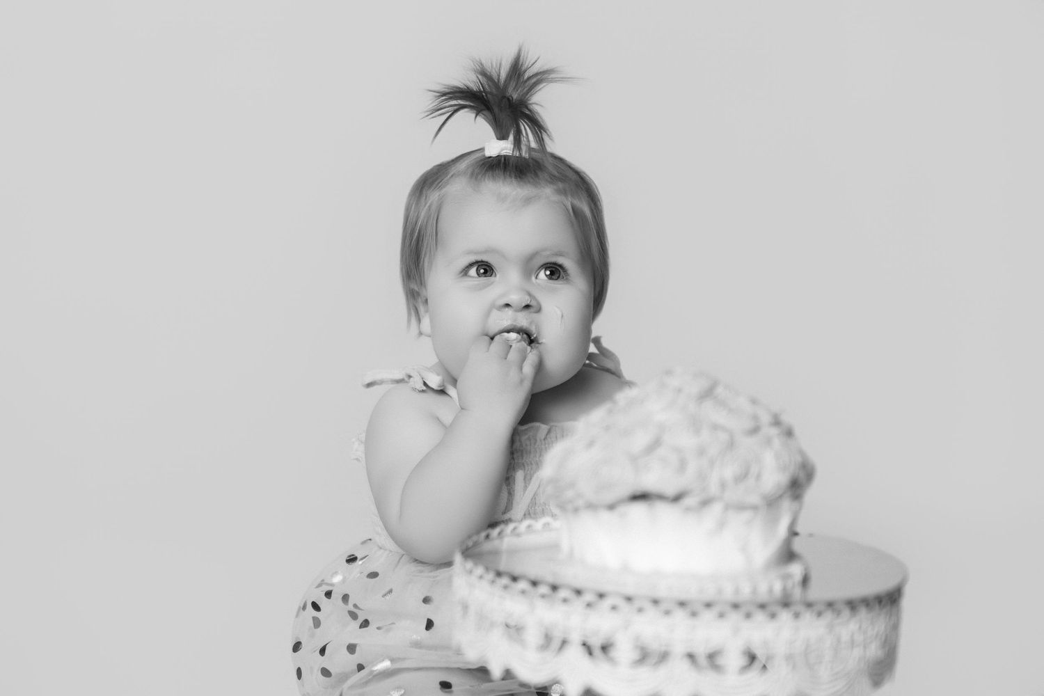 Black and white image of baby exploring cake on decorative stand during first birthday photoshoot.