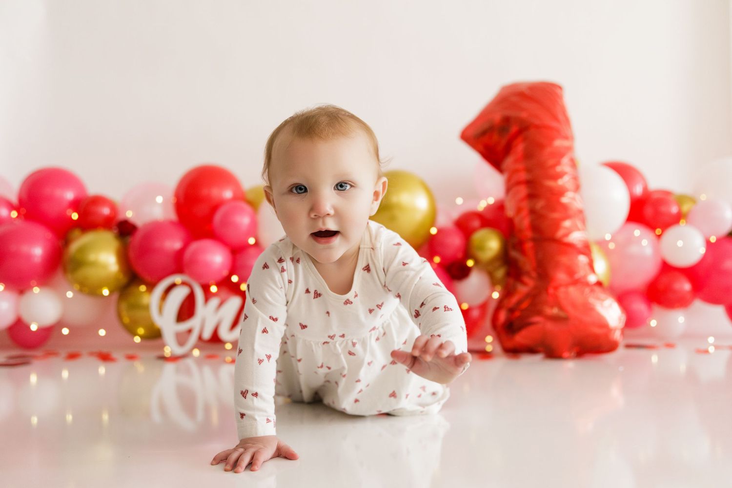 Baby in white patterned outfit crawls on floor with red and gold balloon decorations and number one backdrop.