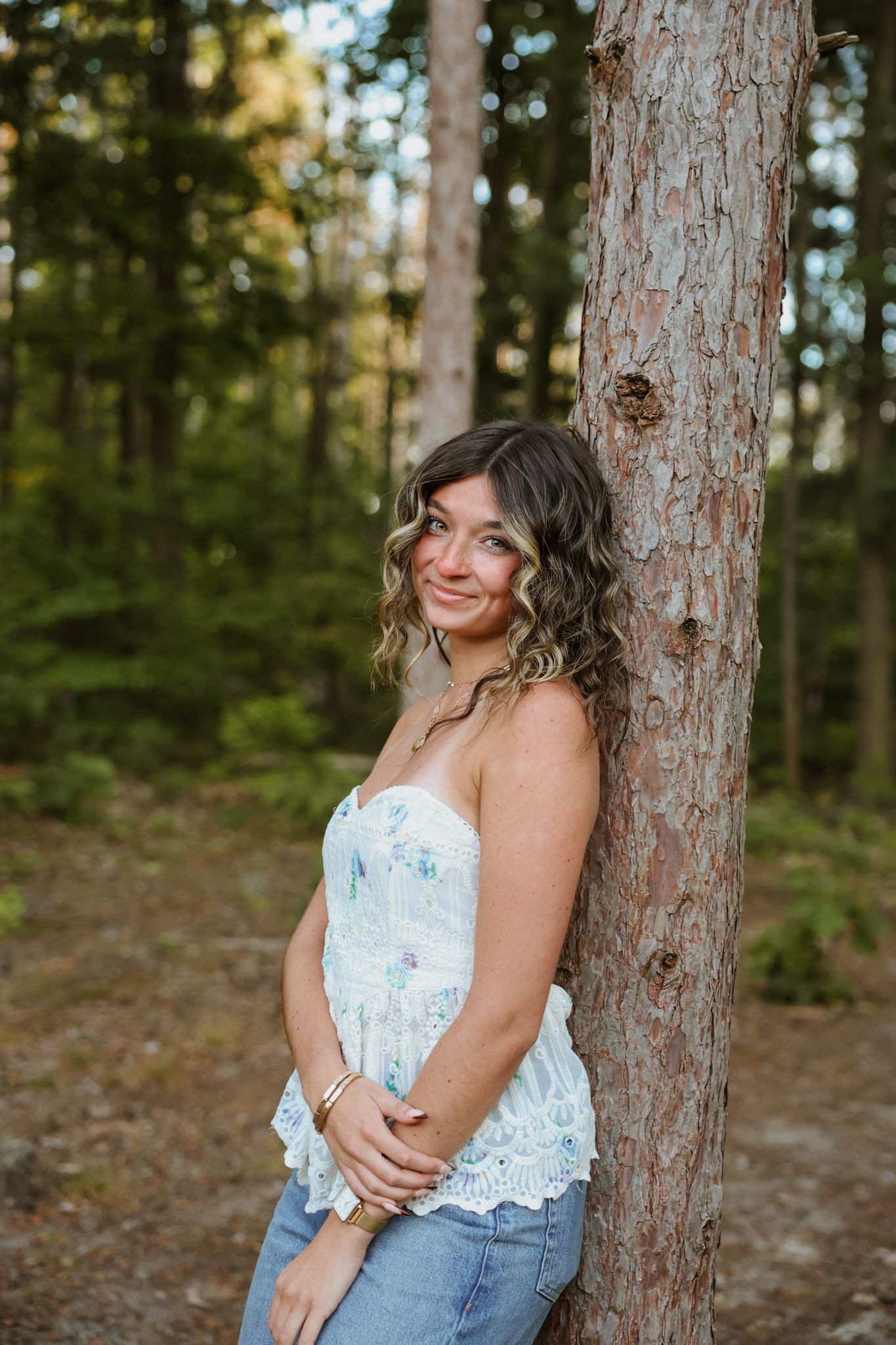 A person in a white lace top and jeans poses against pine trees in a forest setting during a photo session.