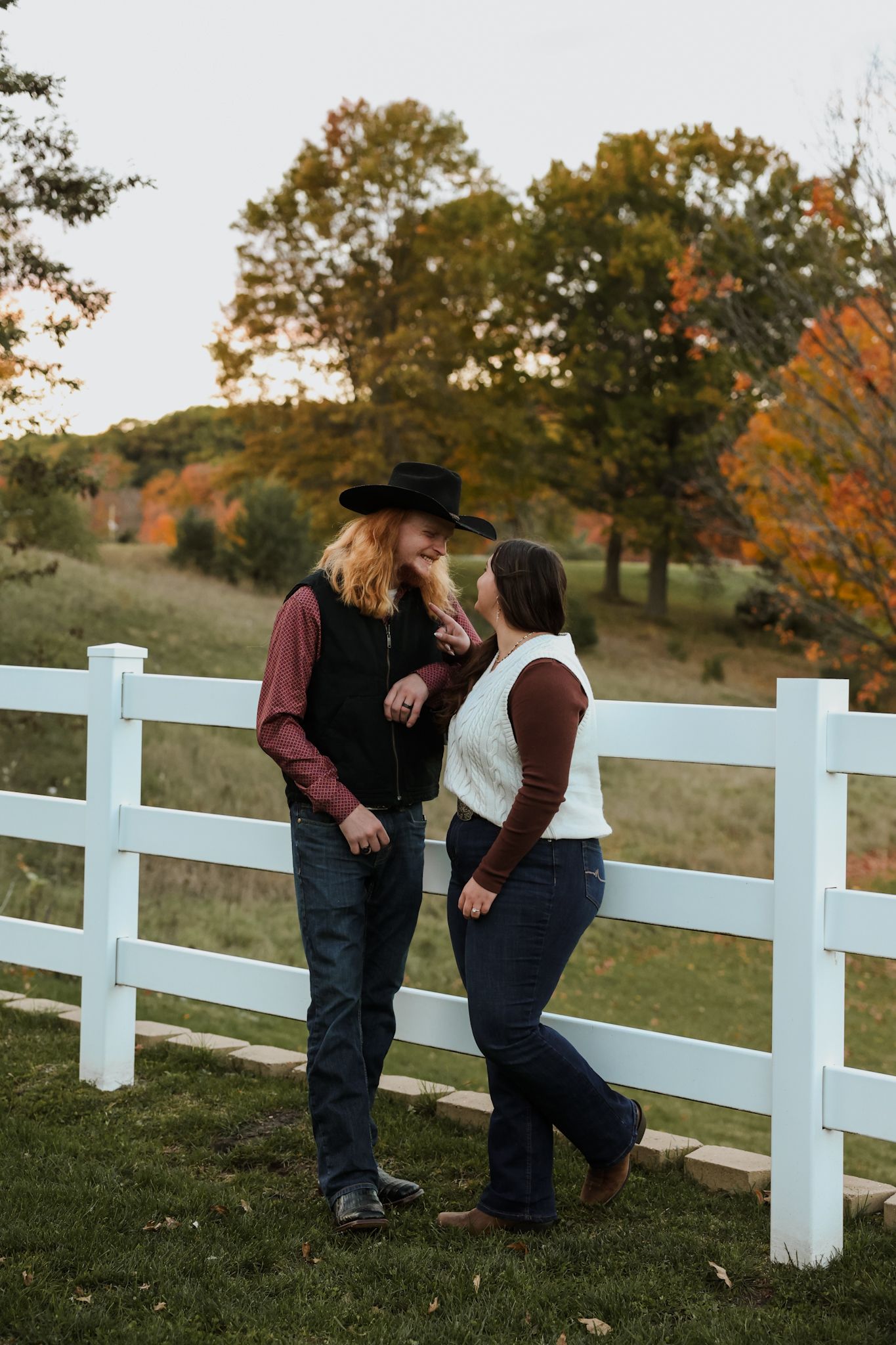 Couple in cowboy hats leaning against white fence with fall foliage trees in background on a farm.
