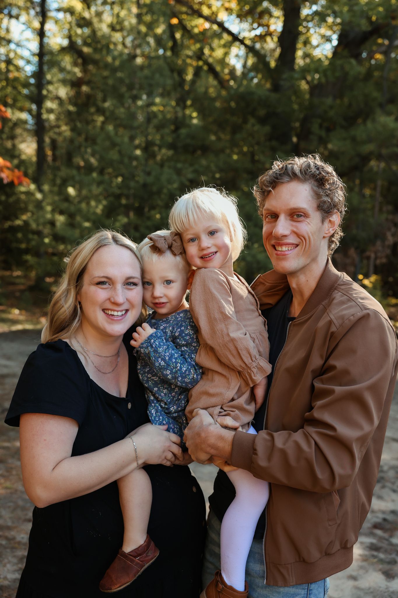 A family of four poses together outdoors in a wooded setting wearing casual clothing.