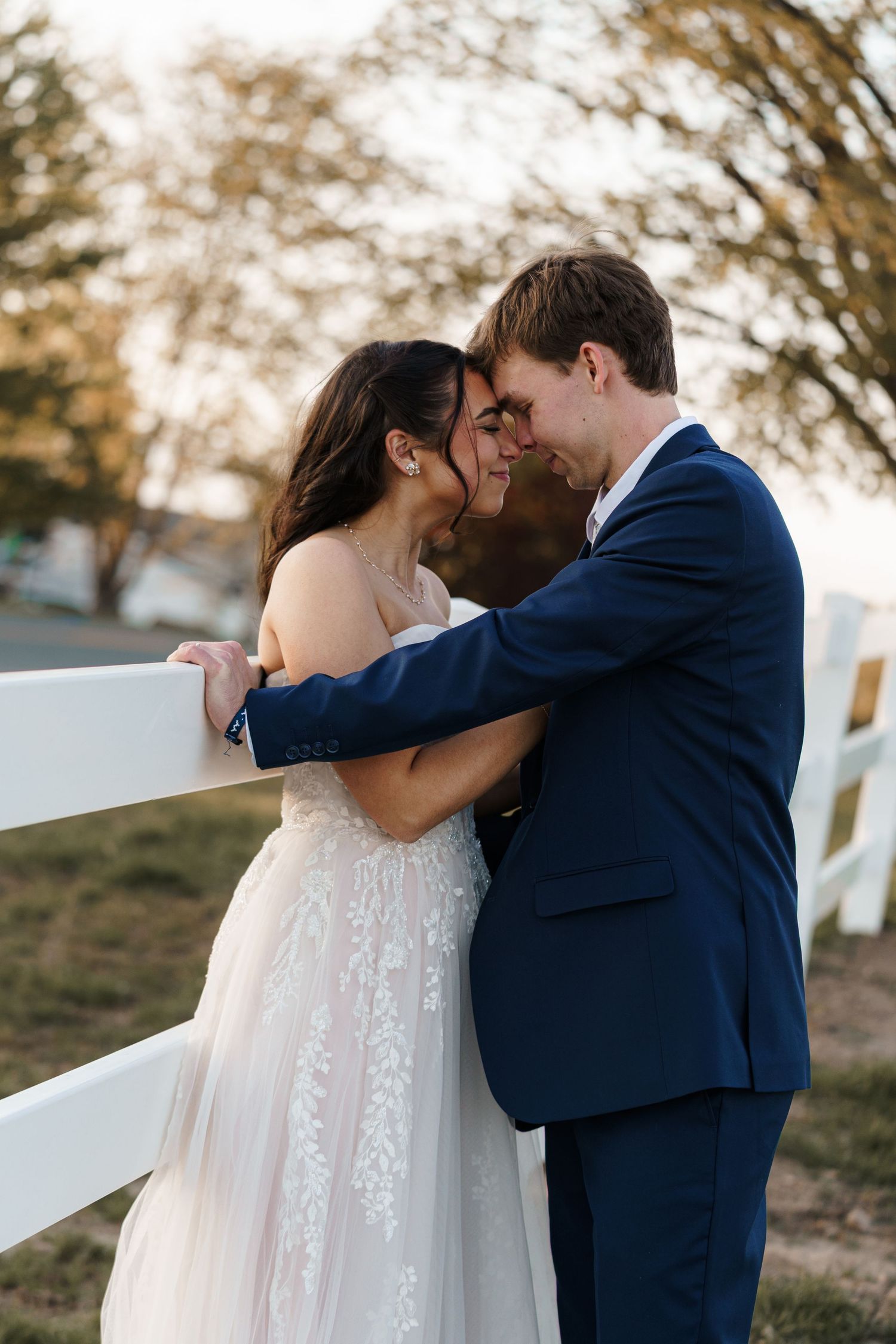 Bride and groom share a romantic moment leaning against a white fence outdoors in wedding attire.