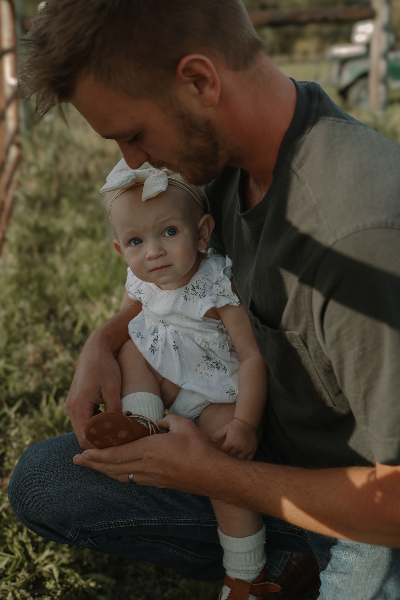 Parent in dark clothing holds small baby wearing white lace dress in outdoor garden setting during sunset.