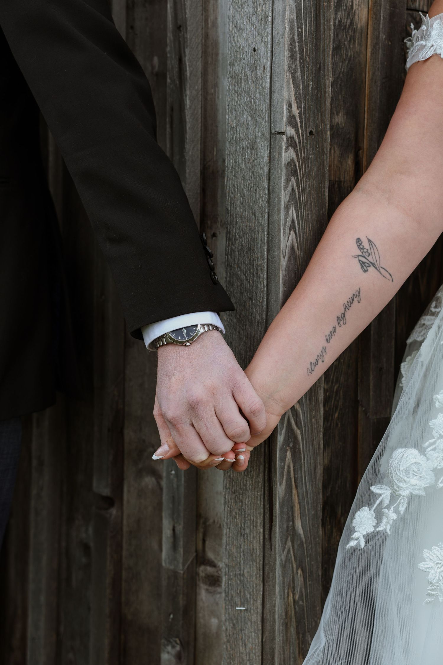 Bride and groom holding hands against rustic wood wall, bride wearing lace dress with forearm tattoo and groom in black suit.