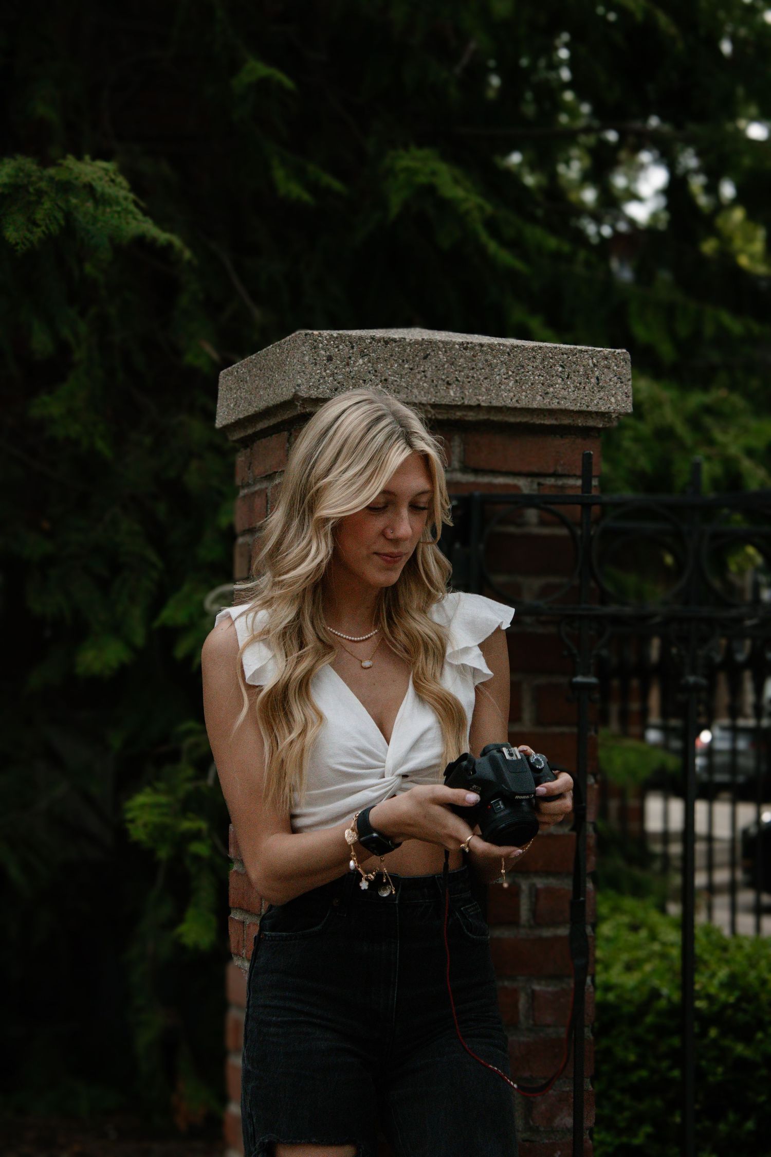 Photographer in white top and dark shorts holds vintage camera near brick pillar in garden setting.