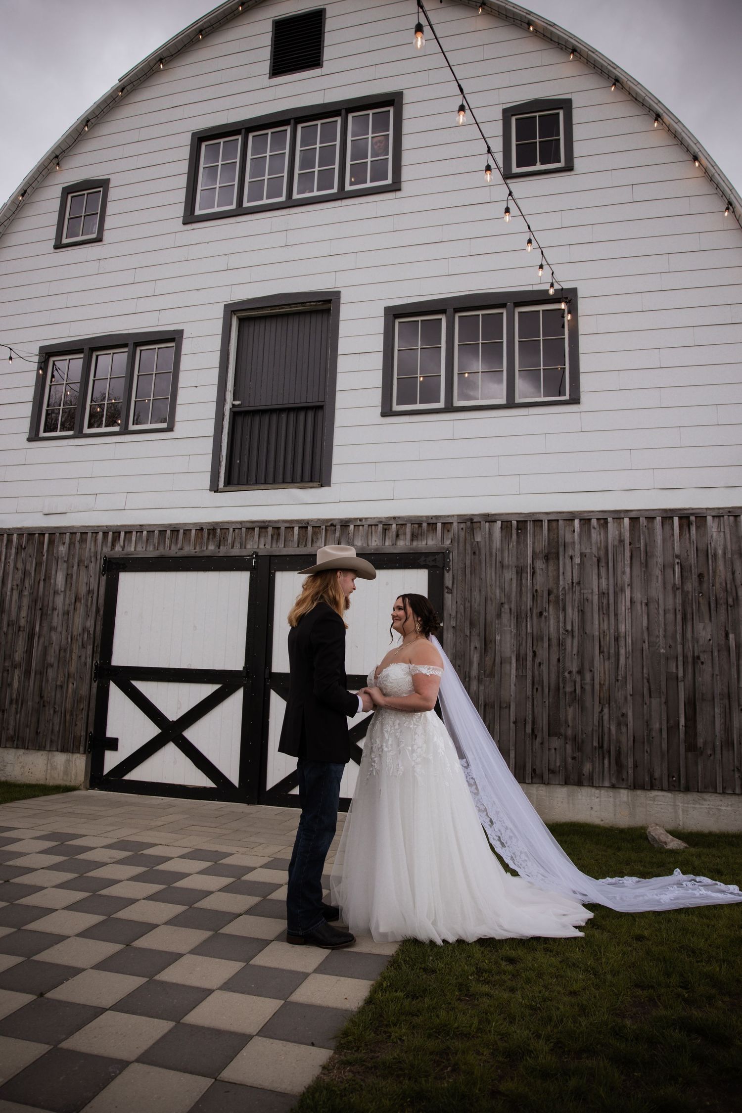 Bride in white gown and veil stands with groom in cowboy hat outside a white barn wedding venue on checkered pavement.
