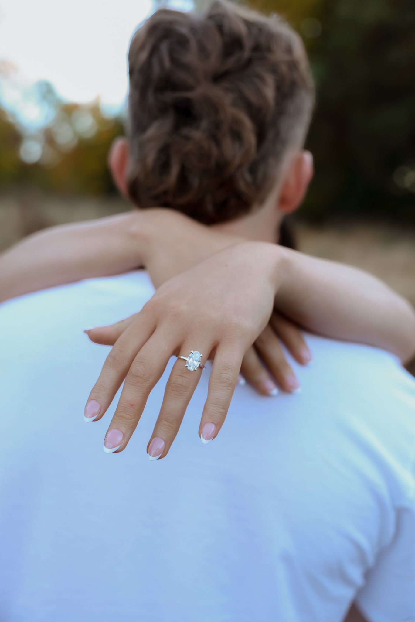 A hand wearing an engagement ring rests on a shoulder while embracing in a romantic moment.