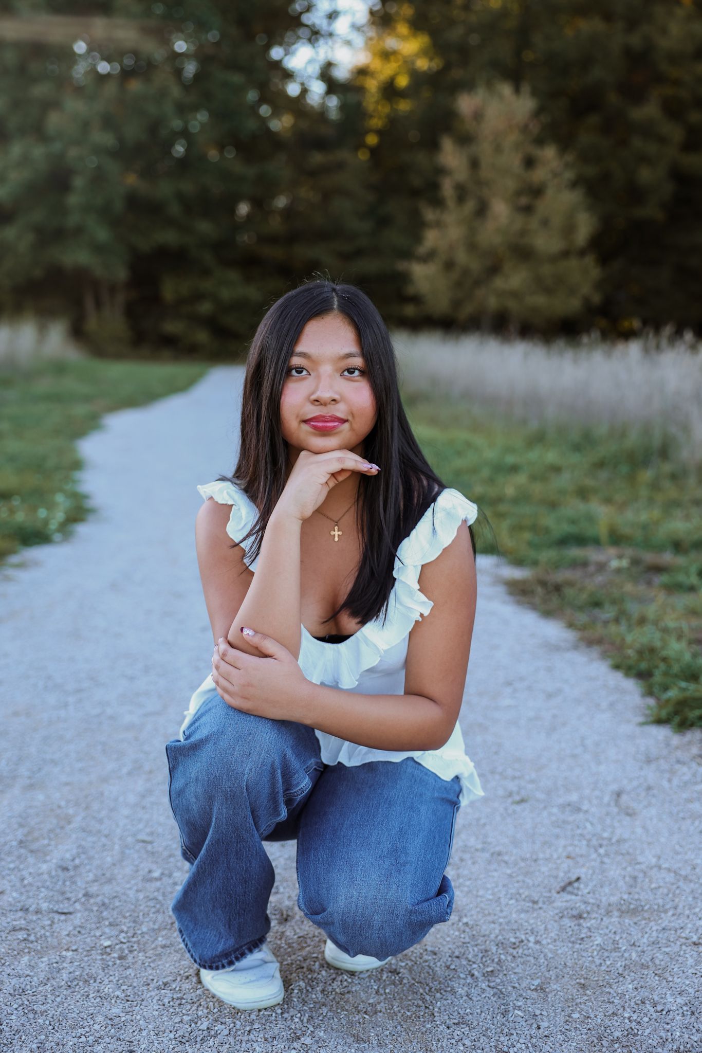 A person in a white ruffled top and blue jeans poses casually on a gravel path surrounded by greenery.