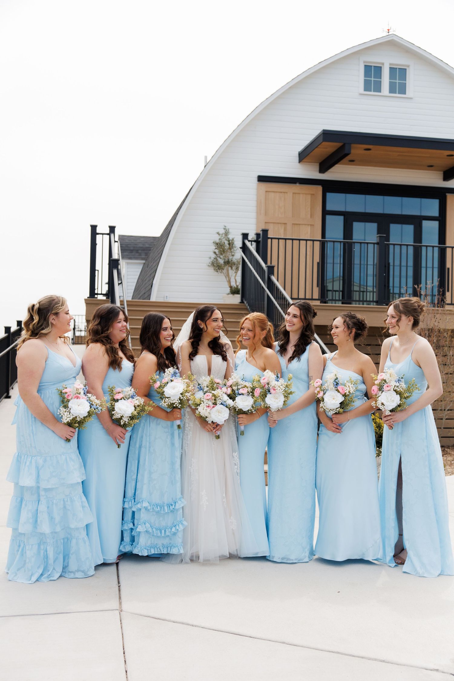 Bride in white gown with six bridesmaids in light blue dresses holding floral bouquets outside a white barn venue.