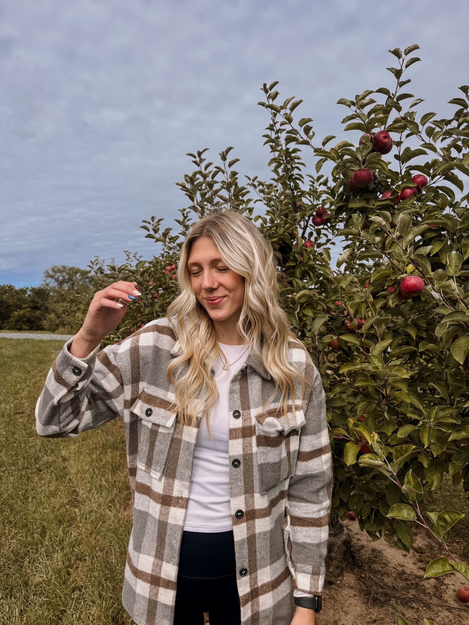 A person wearing a brown plaid shacket and white top stands in front of apple trees during autumn harvest season.