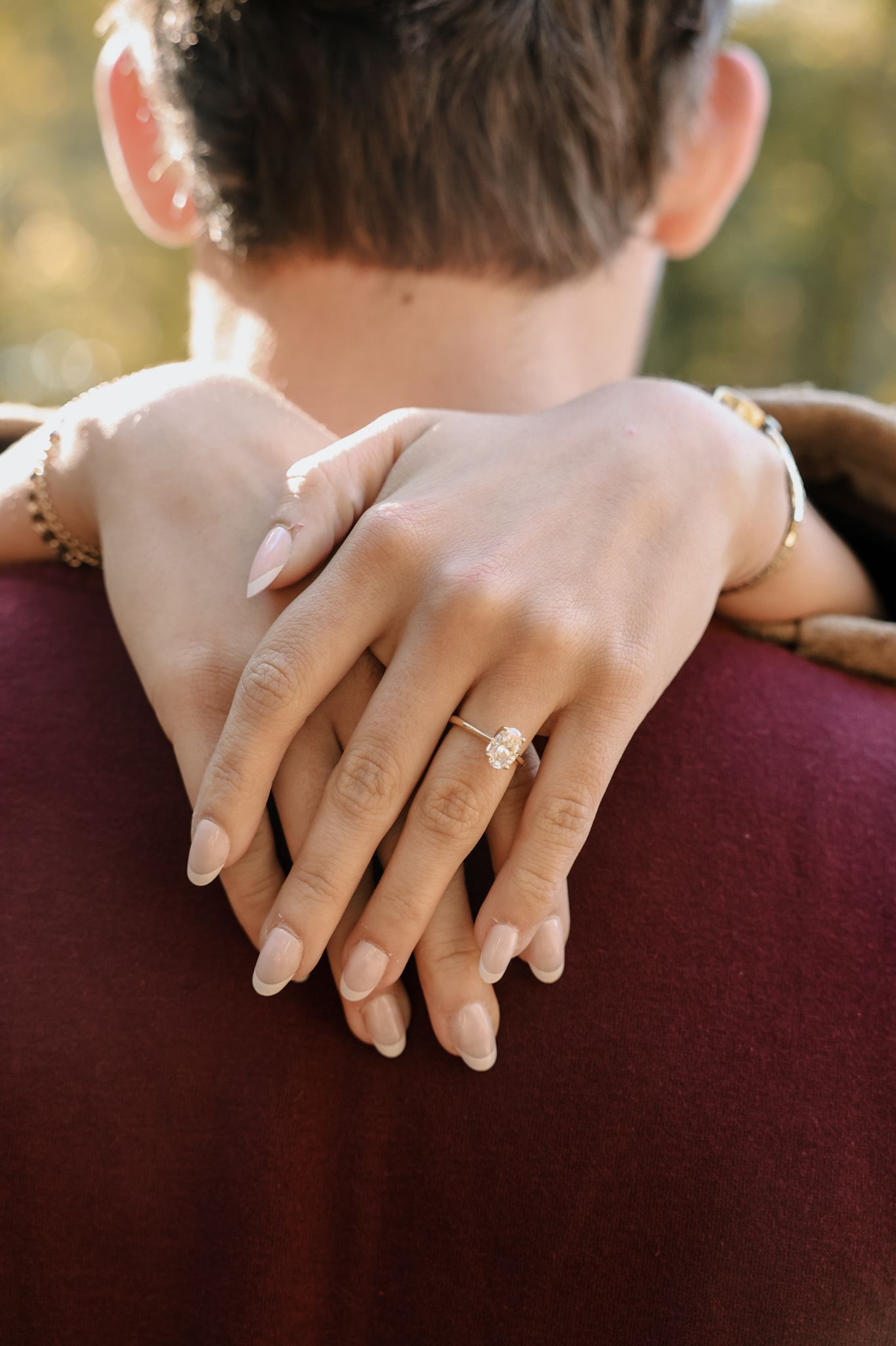 Close-up of hands with engagement ring wrapped around shoulders wearing burgundy sweater in outdoor setting.