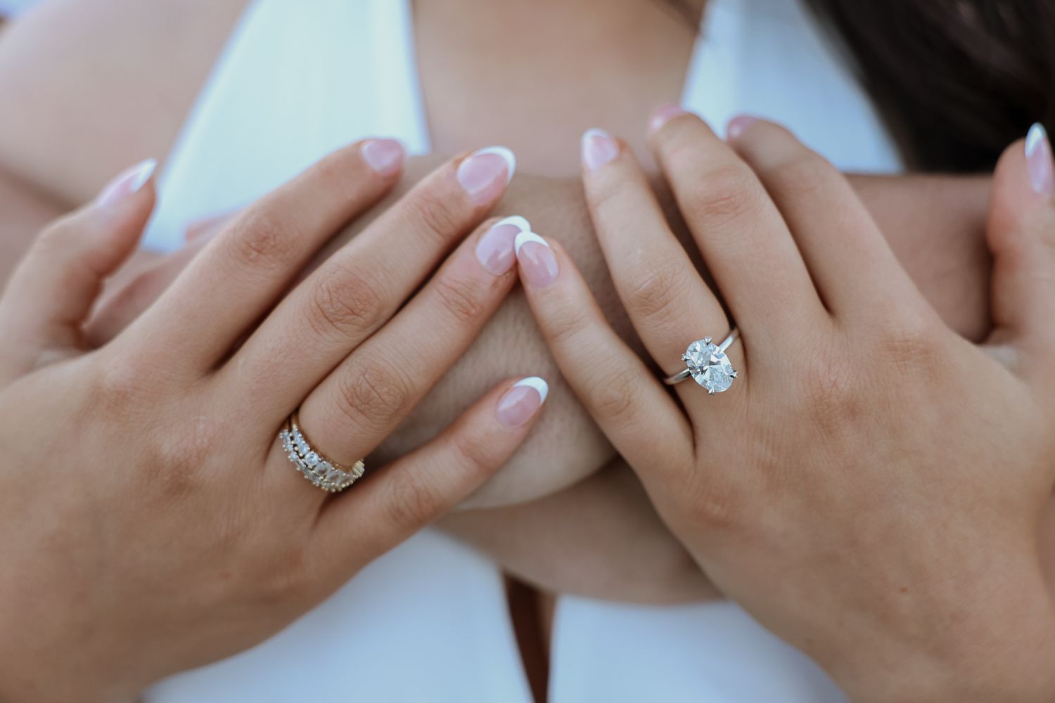 Close-up of two hands displaying elegant diamond engagement rings with French manicured nails on a white background.