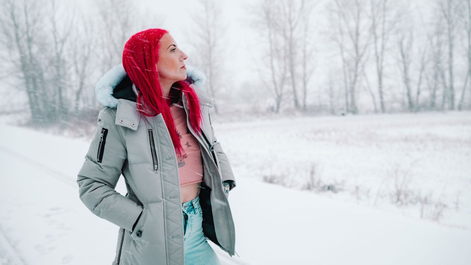 Person wearing a red hat and gray winter coat stands in a snowy winter landscape looking up at the sky.