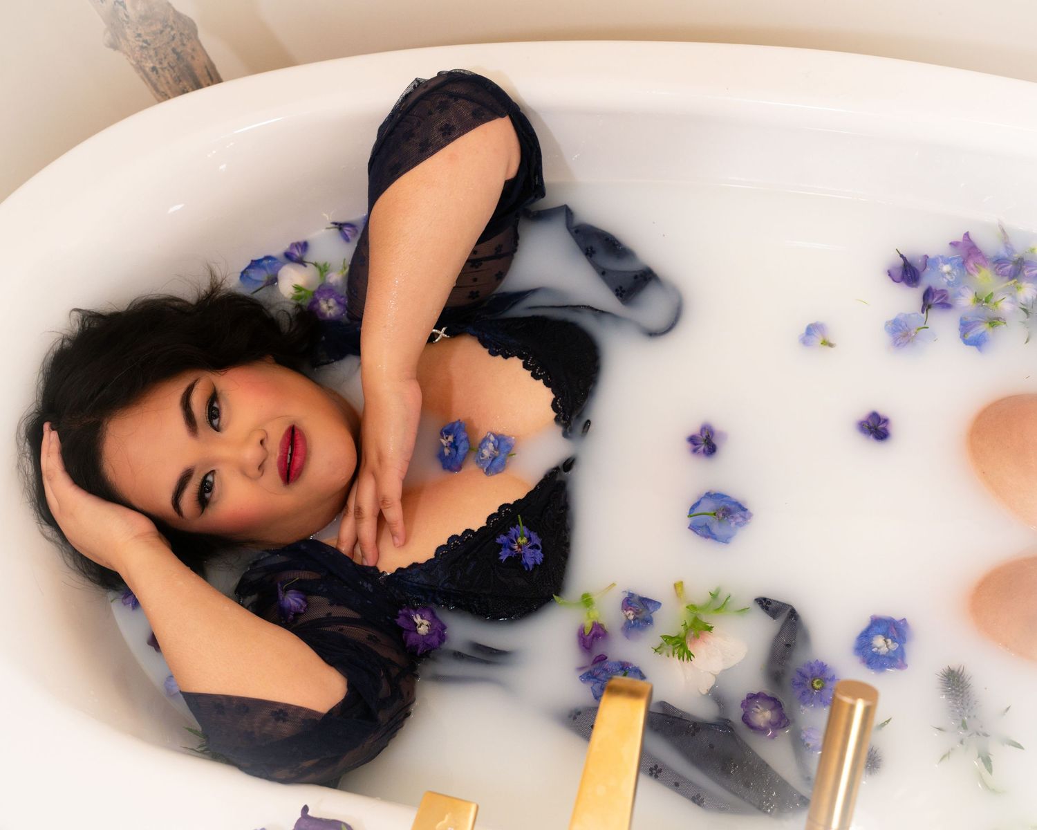 Artistic bathtub portrait with scattered blue and purple flower petals in milky water.