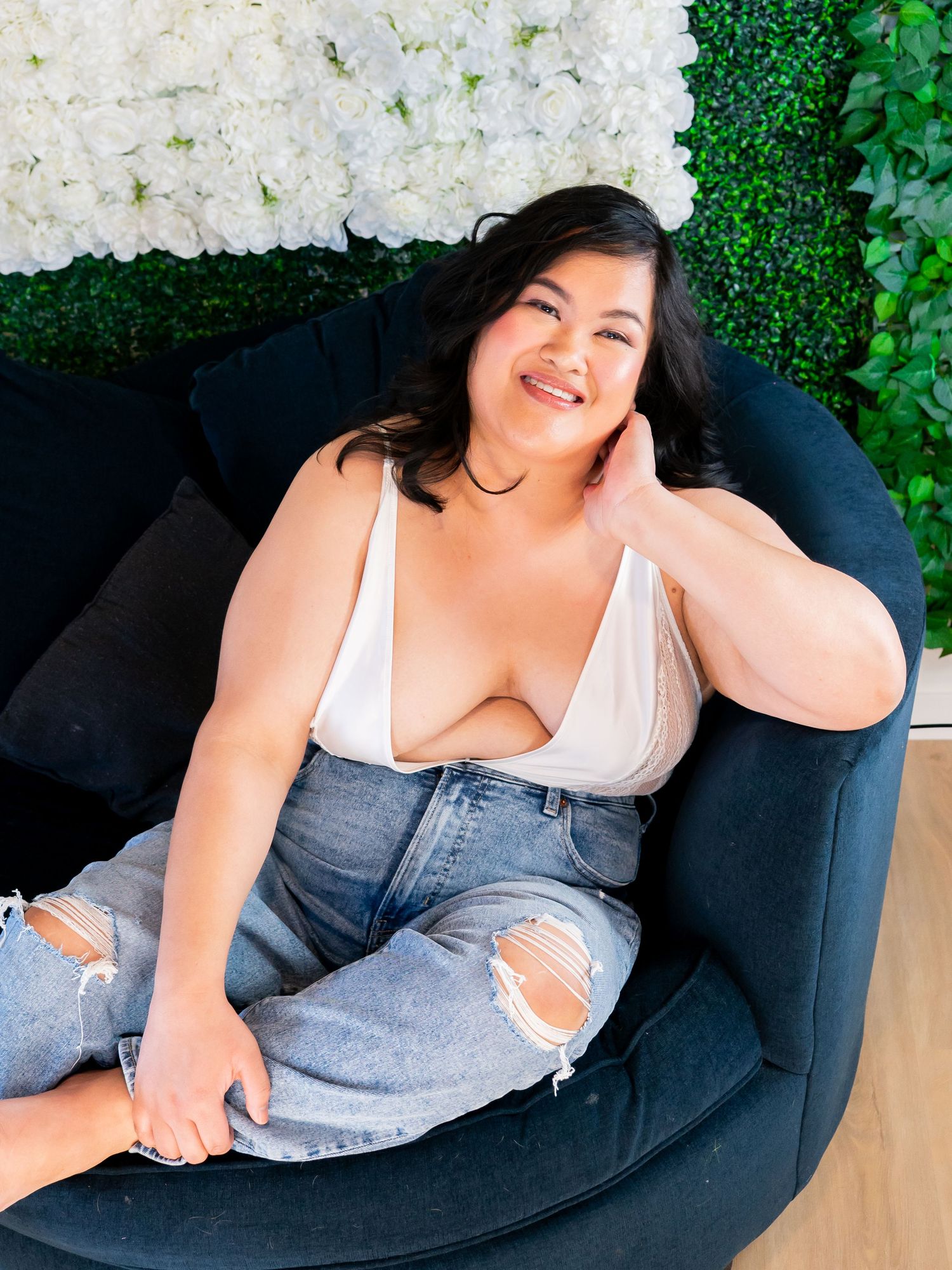 A woman in a white top and distressed jeans lounges comfortably on a navy blue chair against a floral backdrop.