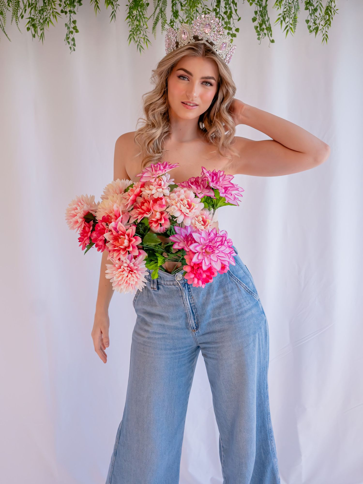 A model wearing wide-leg jeans poses with vibrant pink and coral flowers against a white backdrop with hanging greenery.