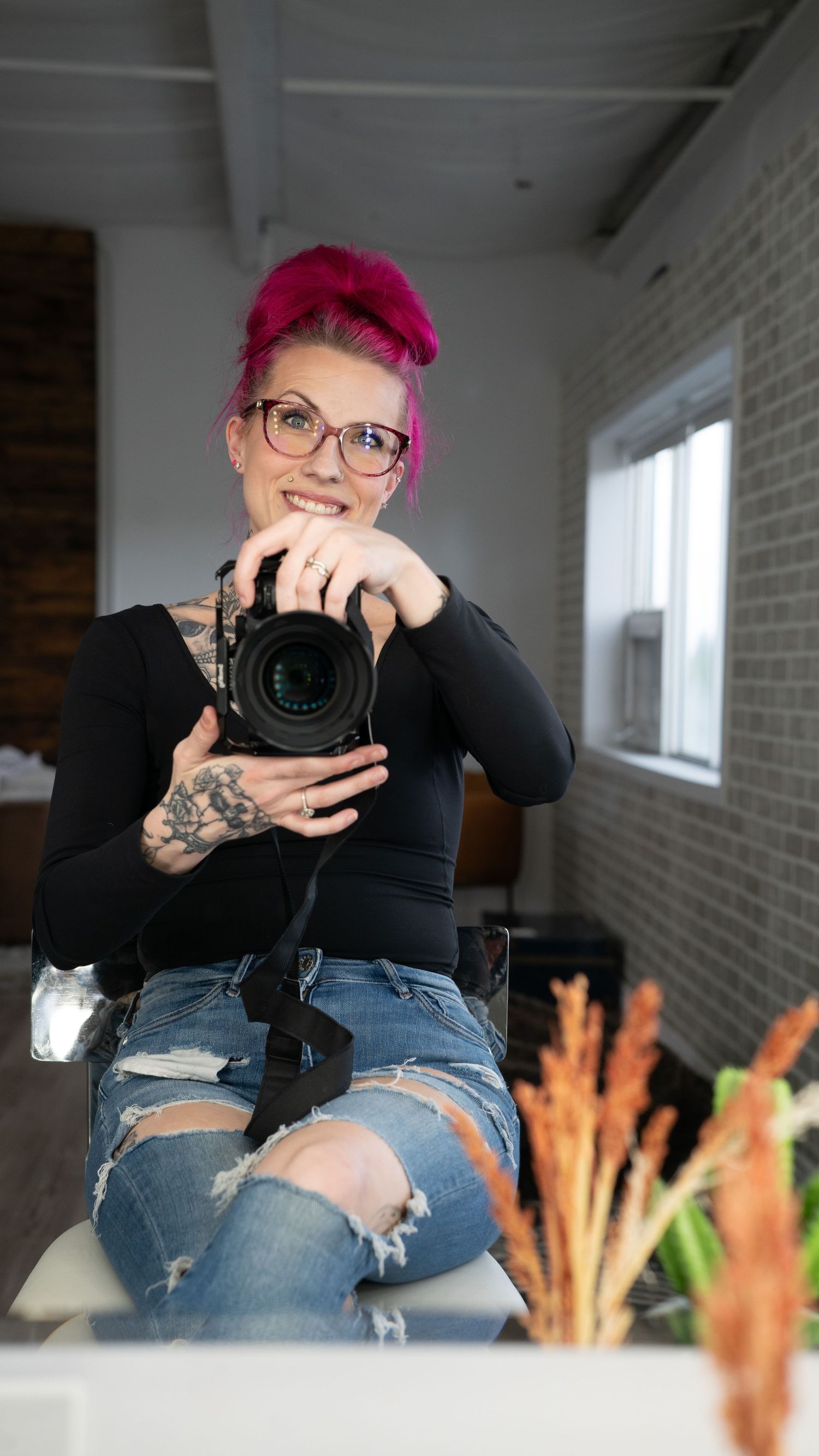 Photographer with bright pink hair and glasses holds professional camera while wearing black top and distressed jeans.