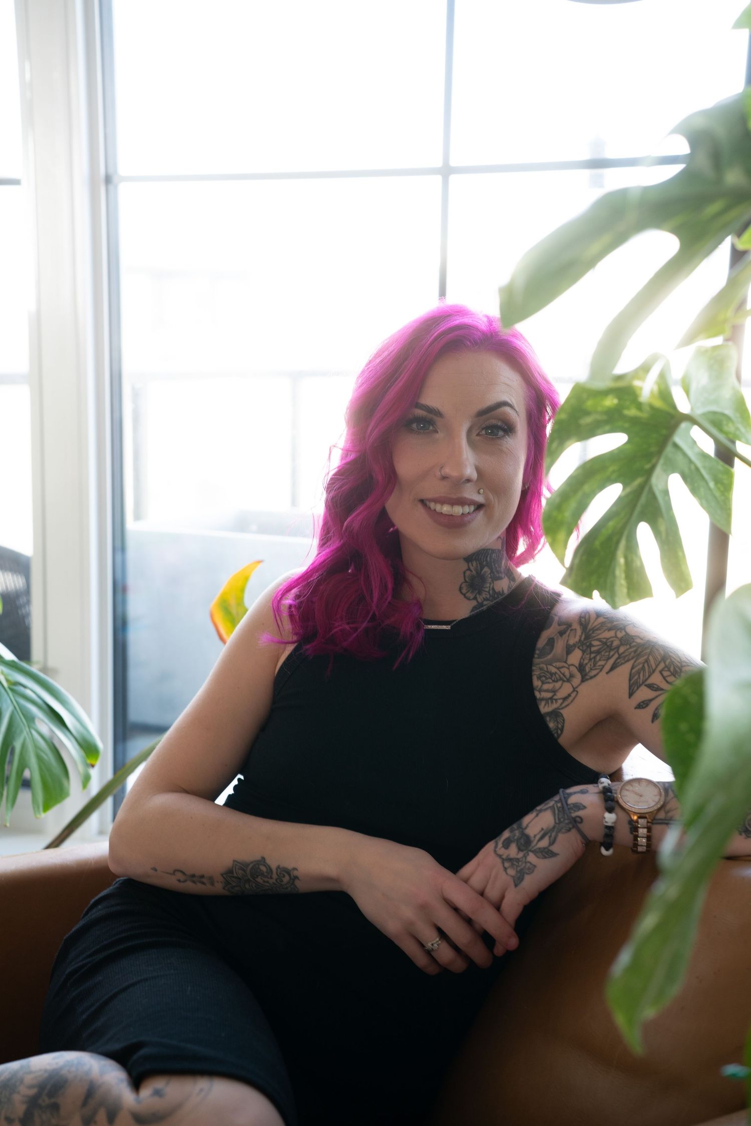 A series of four photos showing a person with bright pink hair and tattoos lounging on a couch near a window with plants.