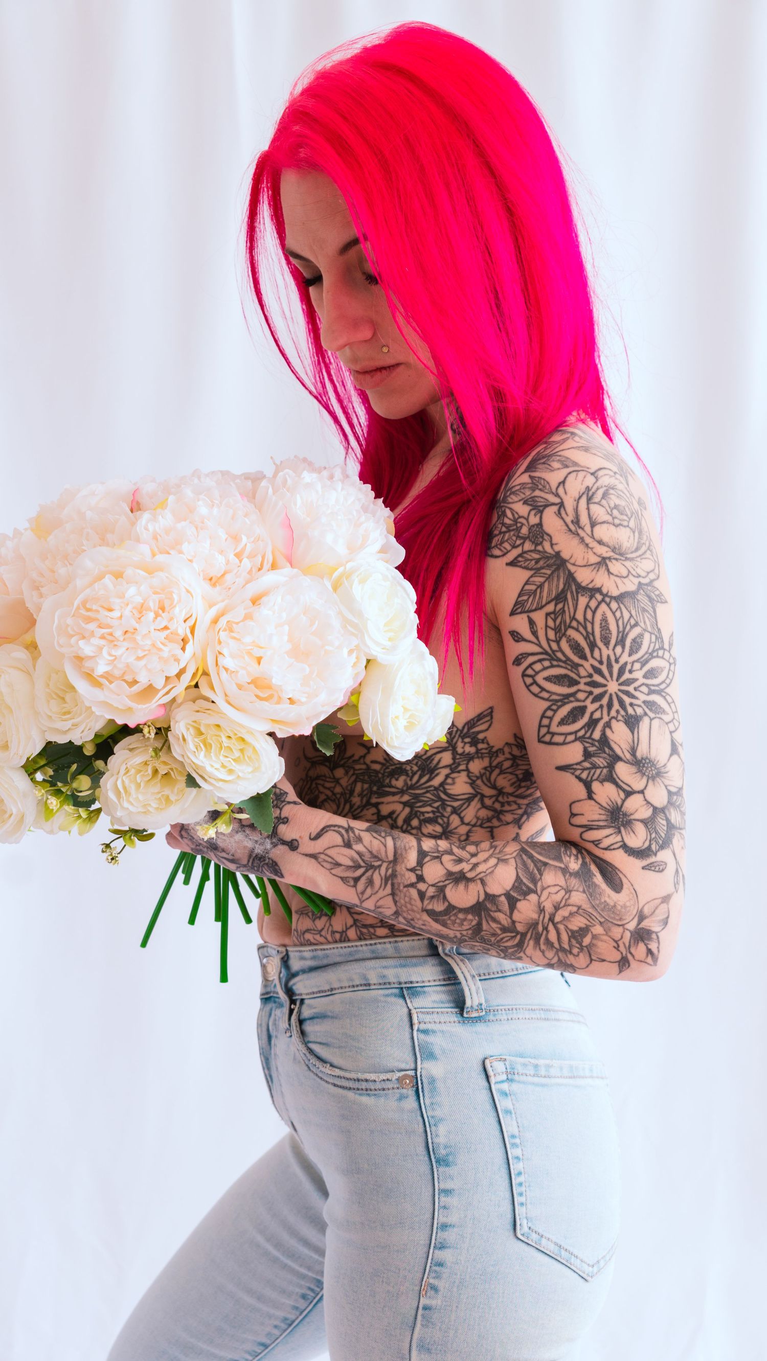 Person with bright pink hair and floral tattoo sleeve holds bouquet of pale pink peonies against white background.