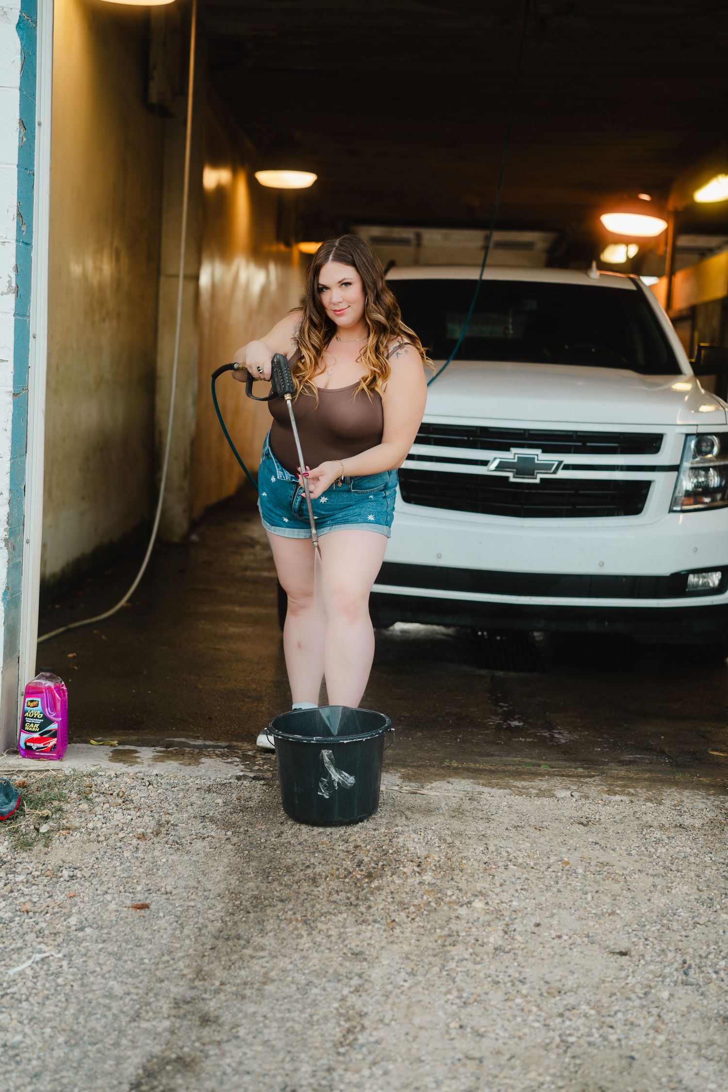 A person in a blue dress crouches down to look under a parked car in a dimly lit parking garage.