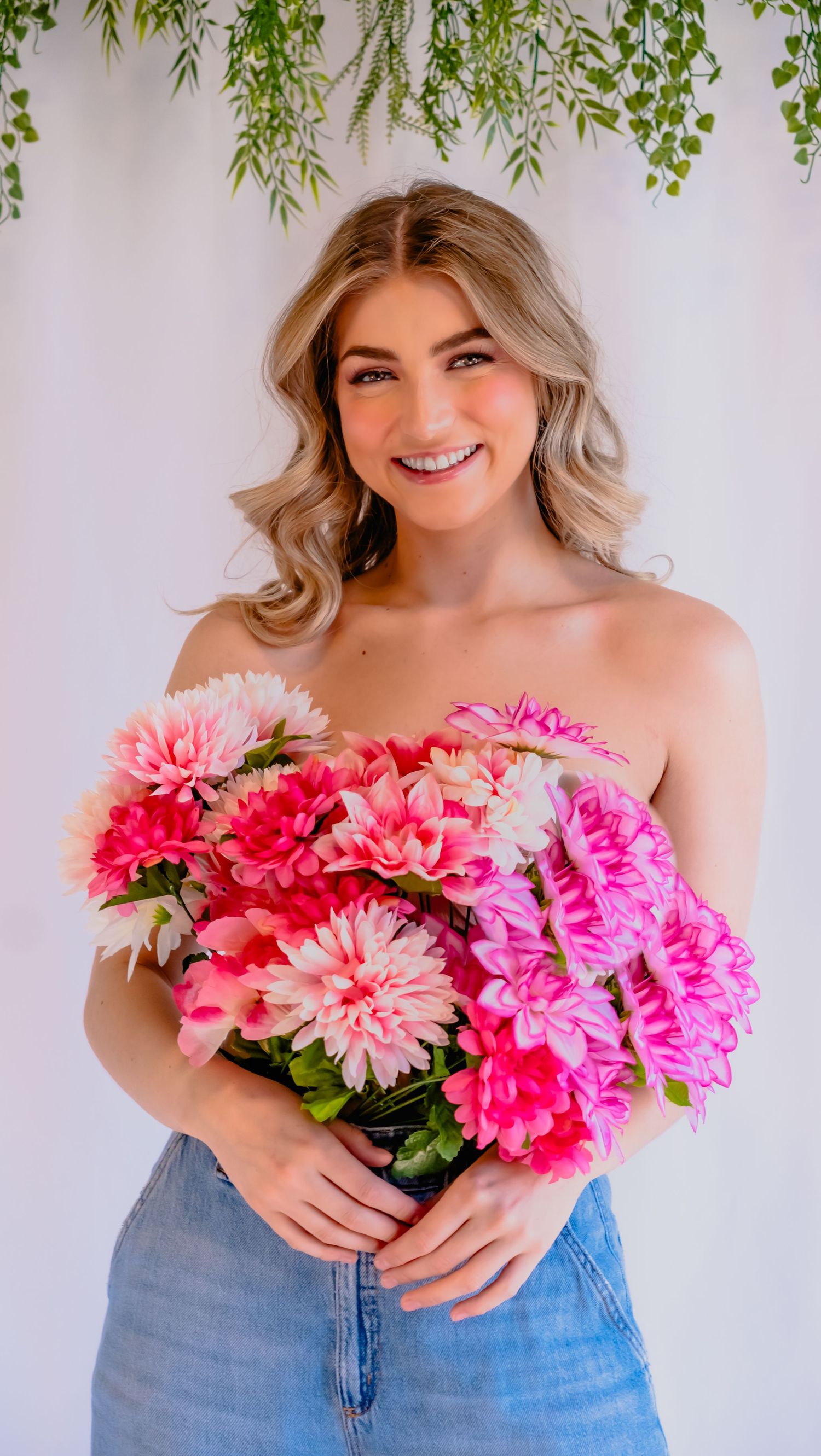 Bright pink and coral dahlia bouquet held against white background with hanging greenery and denim jeans.