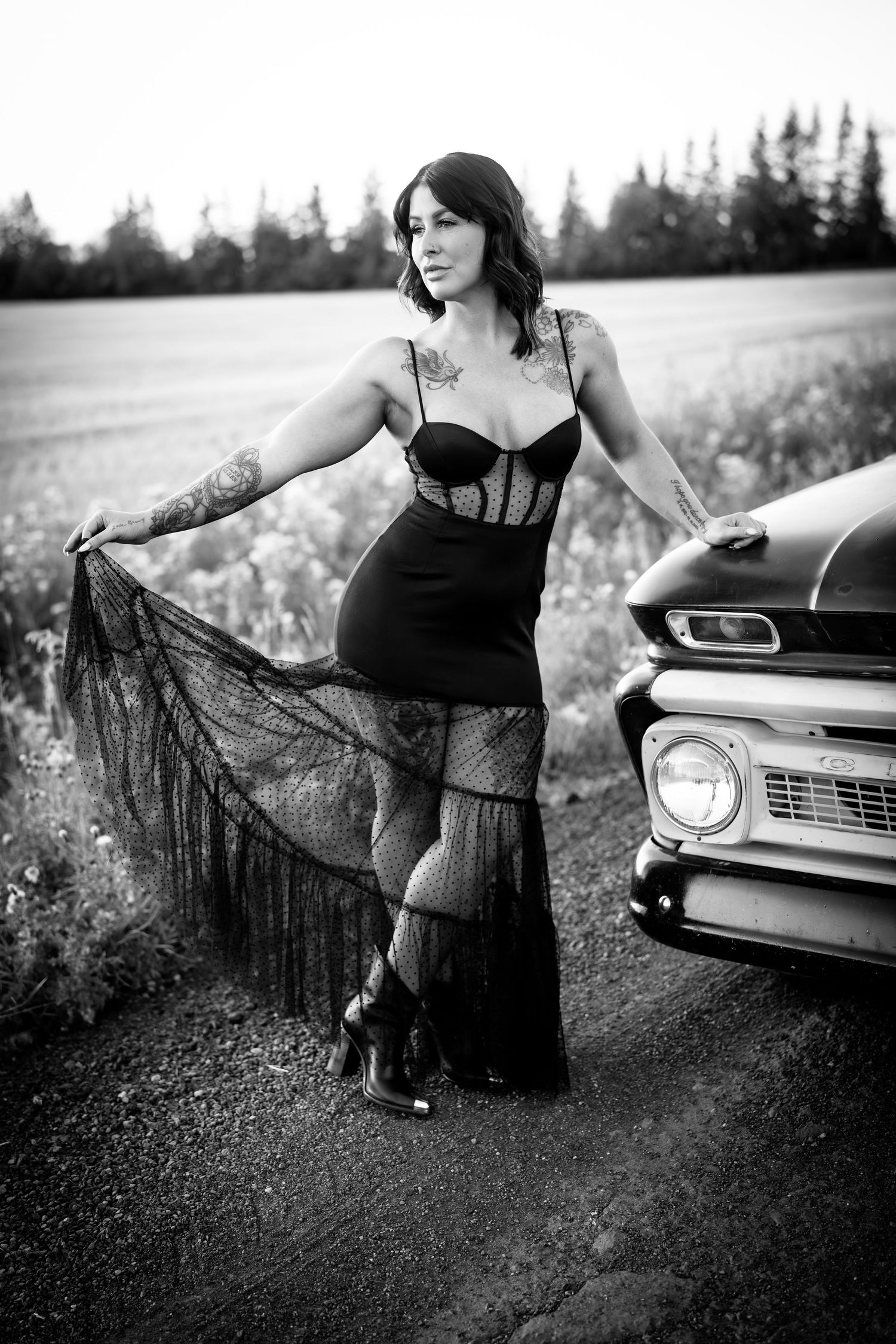 Black and white photo of woman in lace dress posing next to vintage truck in rural field setting.