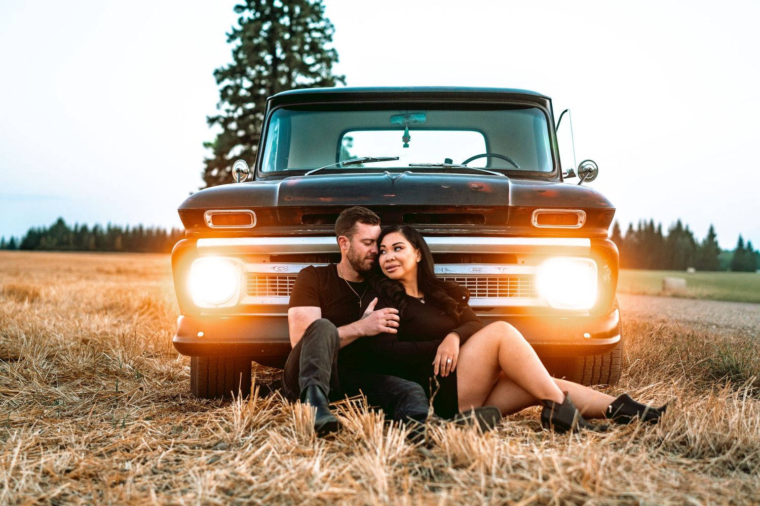 A classic pickup truck sits in a golden field at sunset with its headlights illuminated.