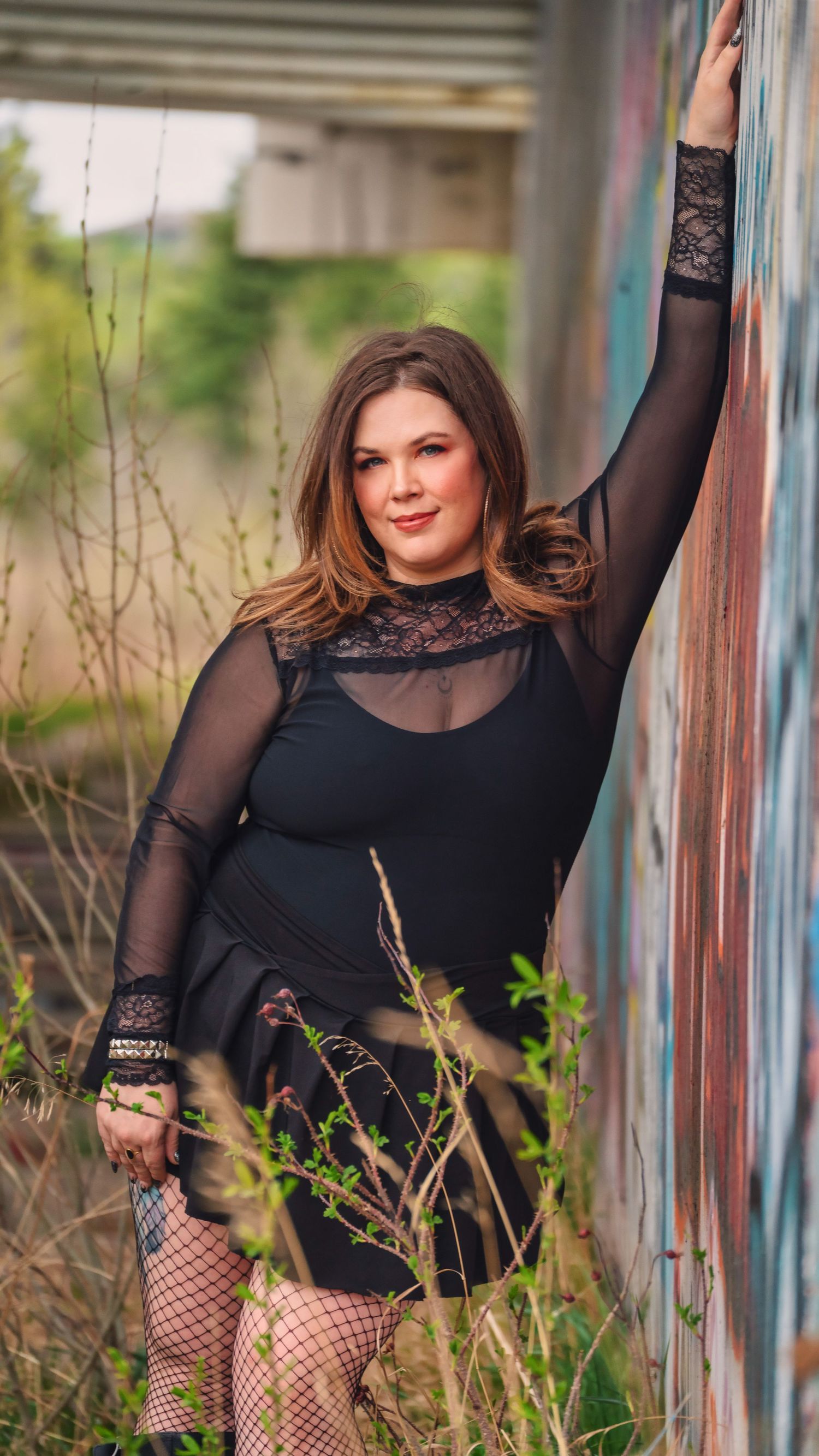 Fashion portrait outdoors showing someone in a black lace top and fishnet stockings posing against a wall in nature.