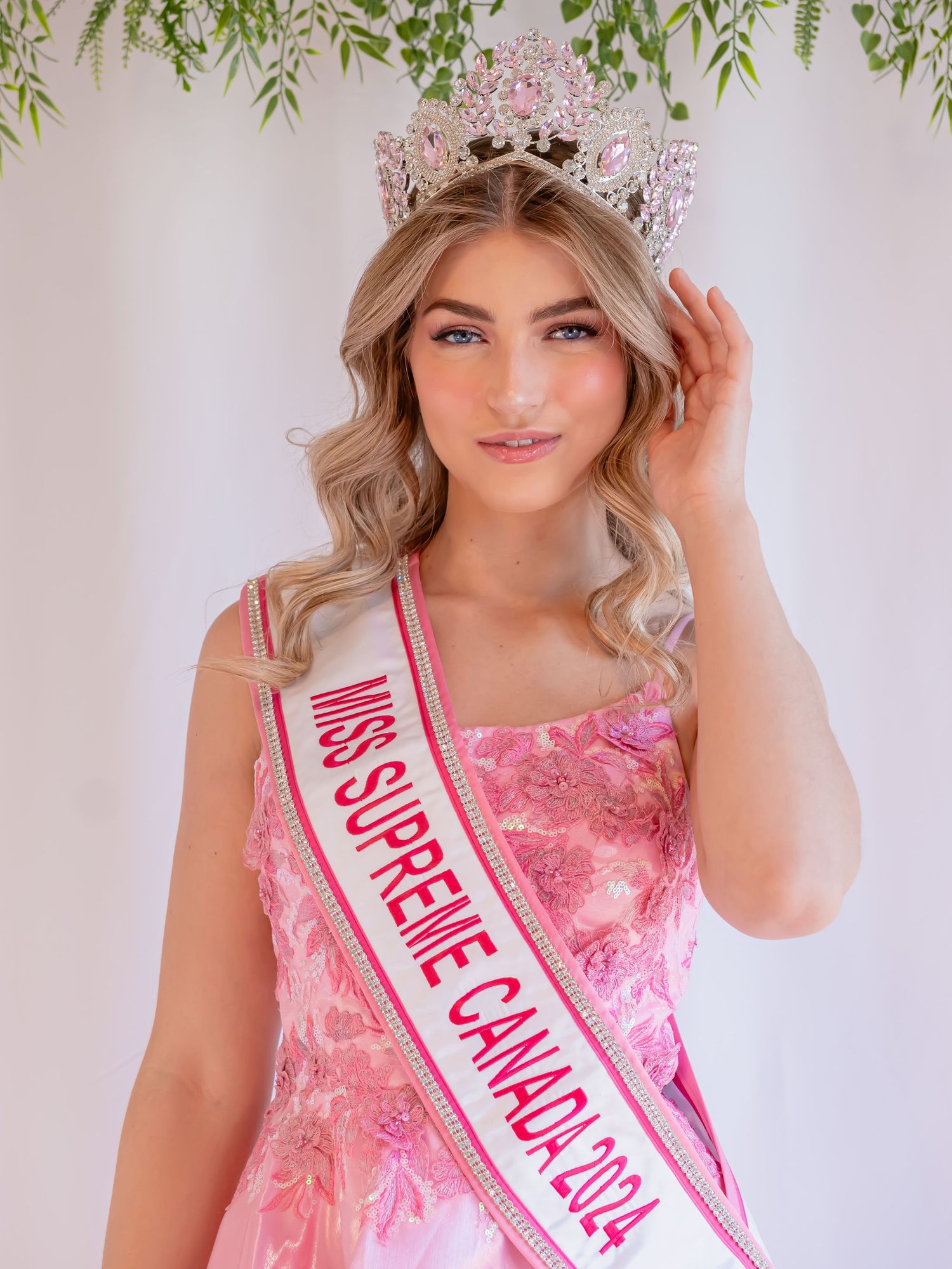 A person wearing a pink floral dress and celebratory sash poses with greenery in the background.
