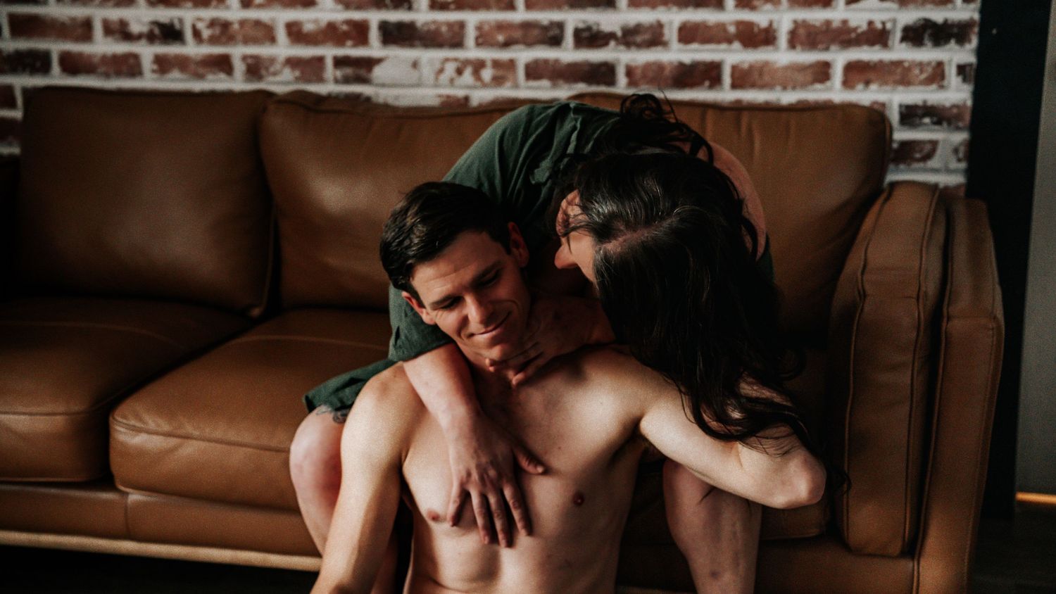 Intimate couple portrait on brown leather couch against exposed brick wall background.