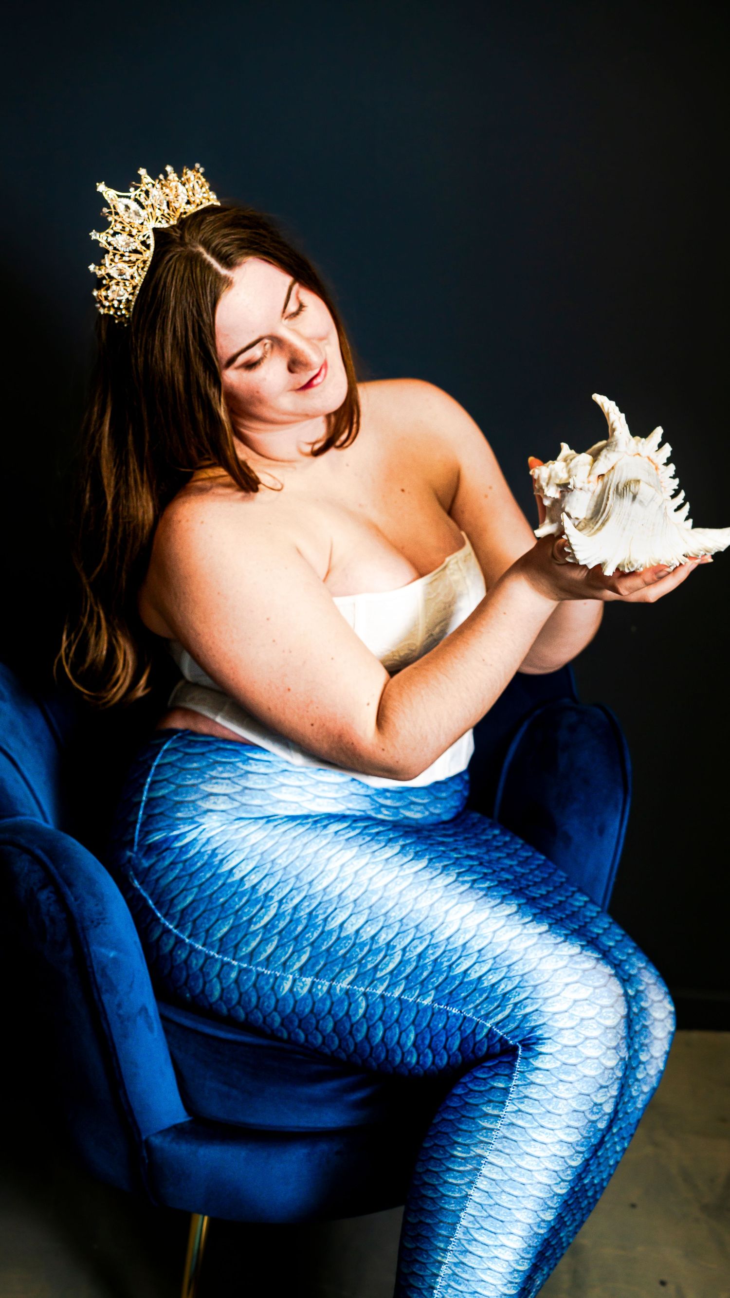 Fantasy mermaid photoshoot featuring blue tail costume, crown, and decorative seashell prop.