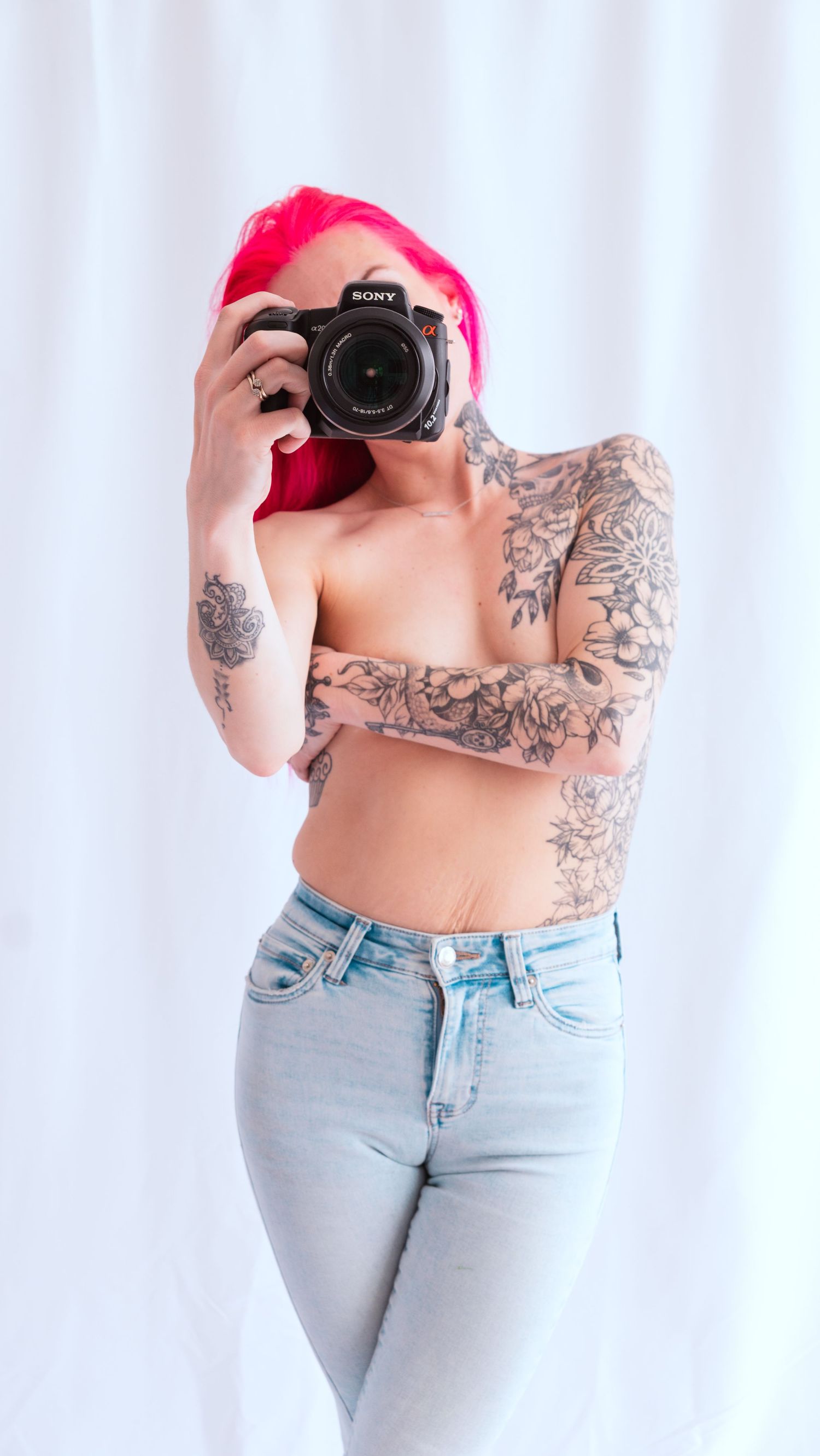 Photographer in light jeans with pink hair and sleeve tattoos holds DSLR camera against white backdrop.