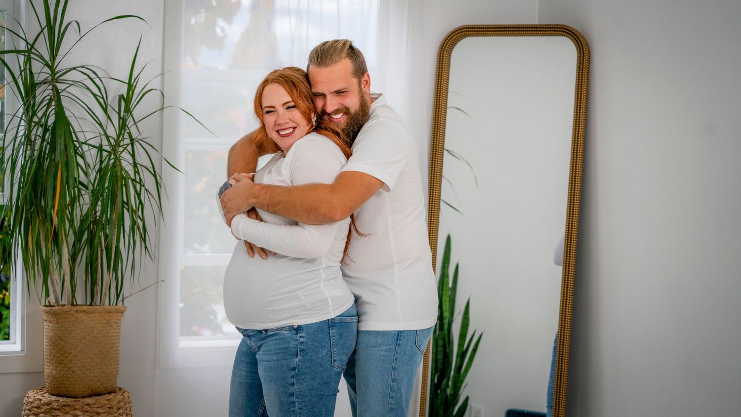 Intimate couple portrait in casual white shirts embracing by mirror and houseplant.