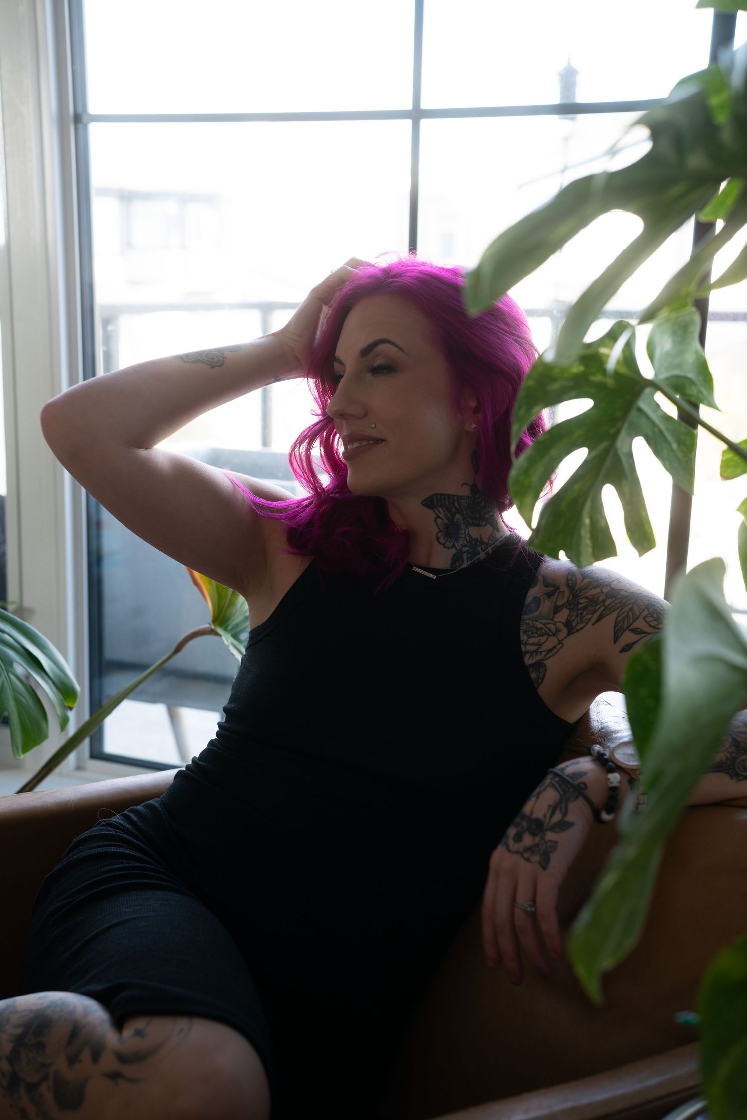 A person with bright pink hair lounges on a brown couch near a window with houseplants in view.