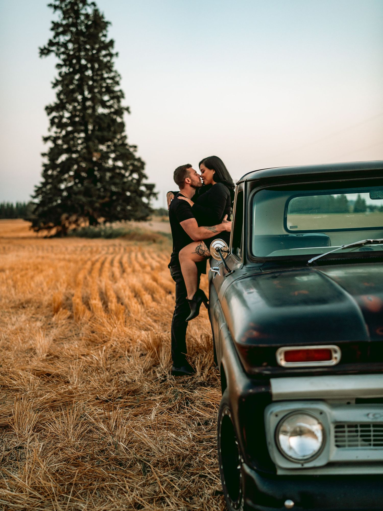 A vintage Chevy pickup truck parked in a golden wheat field at sunset with a couple embracing beside it.