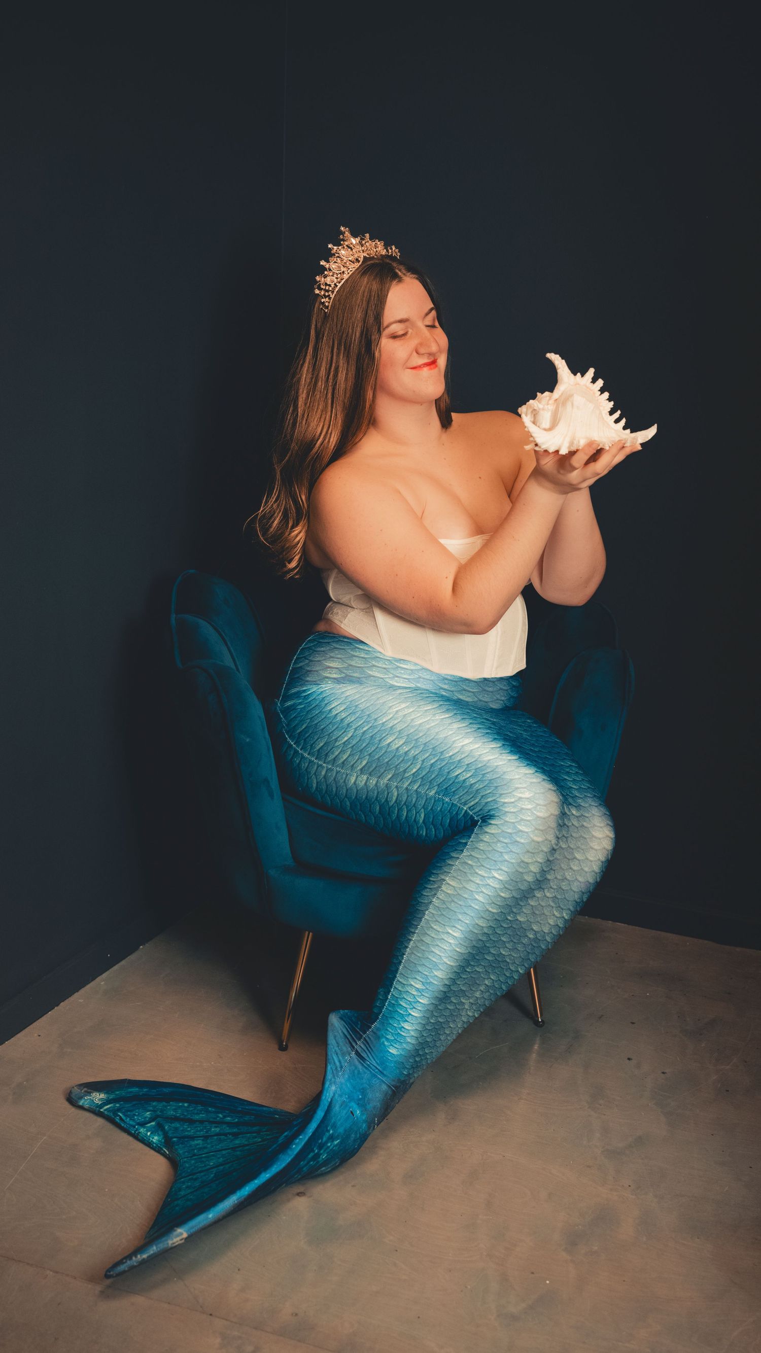 Mermaid-themed portrait with blue tail costume, gold crown and white seashell against dark background.