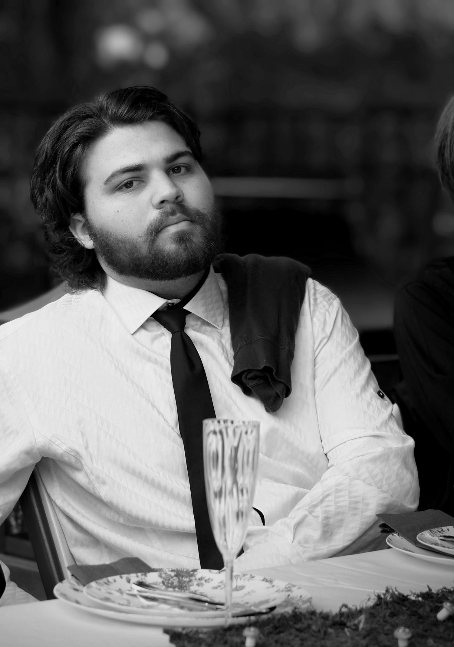 B&W candid senior event portrait—handsome bearded young man in white shirt & tie, seated casually outdoors. Photography by Rachel.