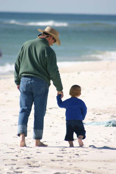 Candid beach portrait of father and toddler son walking hand in hand on white sand at Perdido Key, FL. Photography by Rachel.