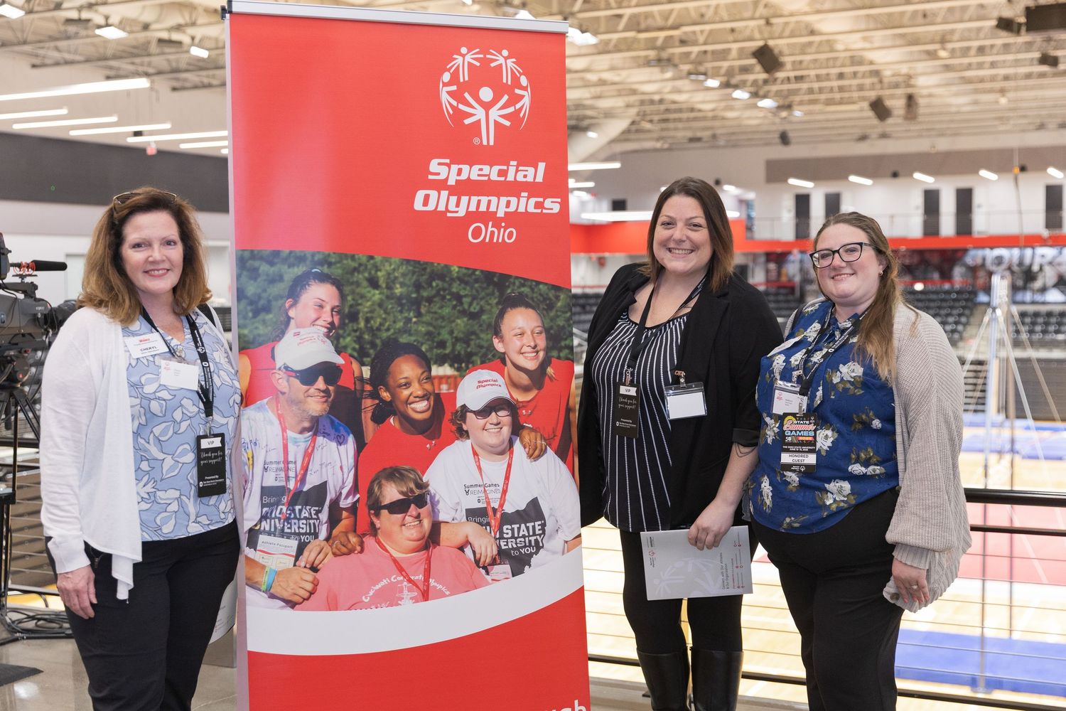 Three women smile beside a Special Olympics Ohio banner at an indoor sporting event.