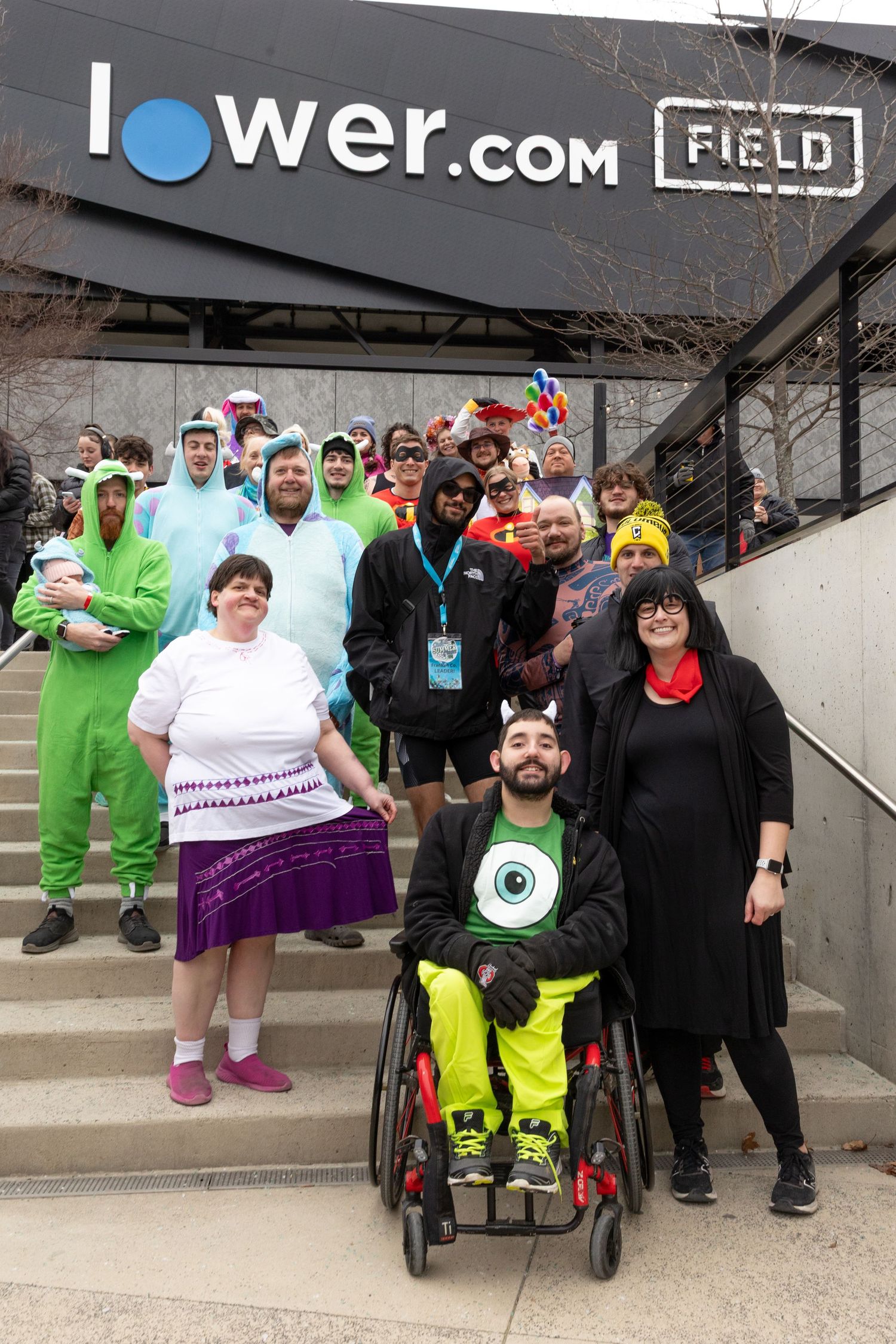 Group in costumes posing outside Lower.com Field, including a person in a wheelchair wearing a Monsters Inc. outfit.
