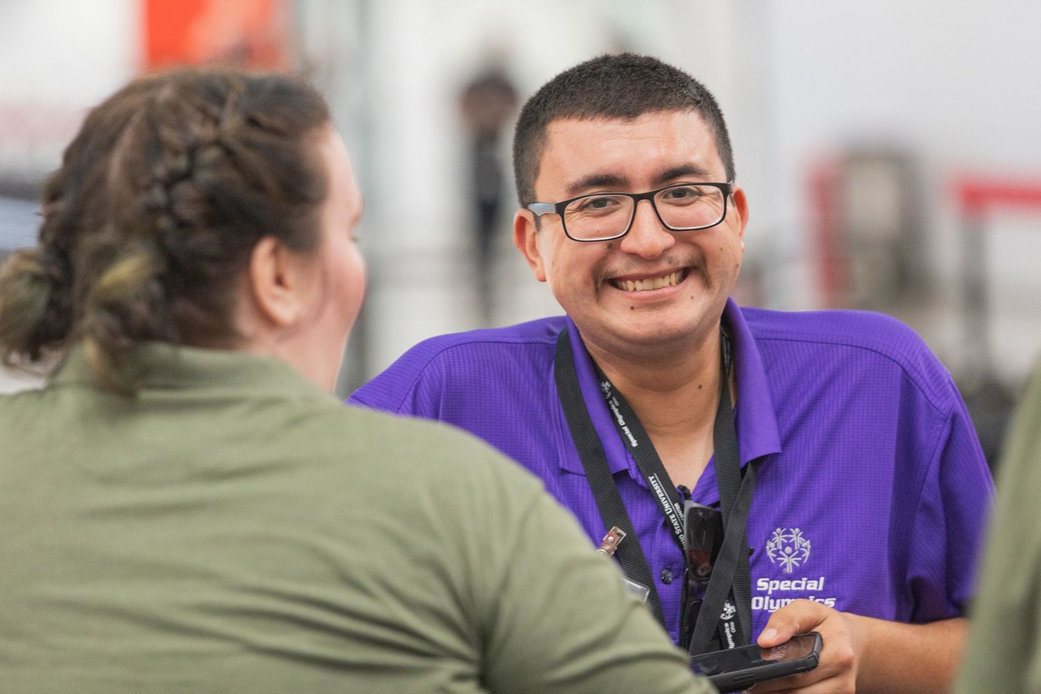 A smiling Special Olympics volunteer in a purple shirt engages warmly with an athlete at an indoor sporting event.